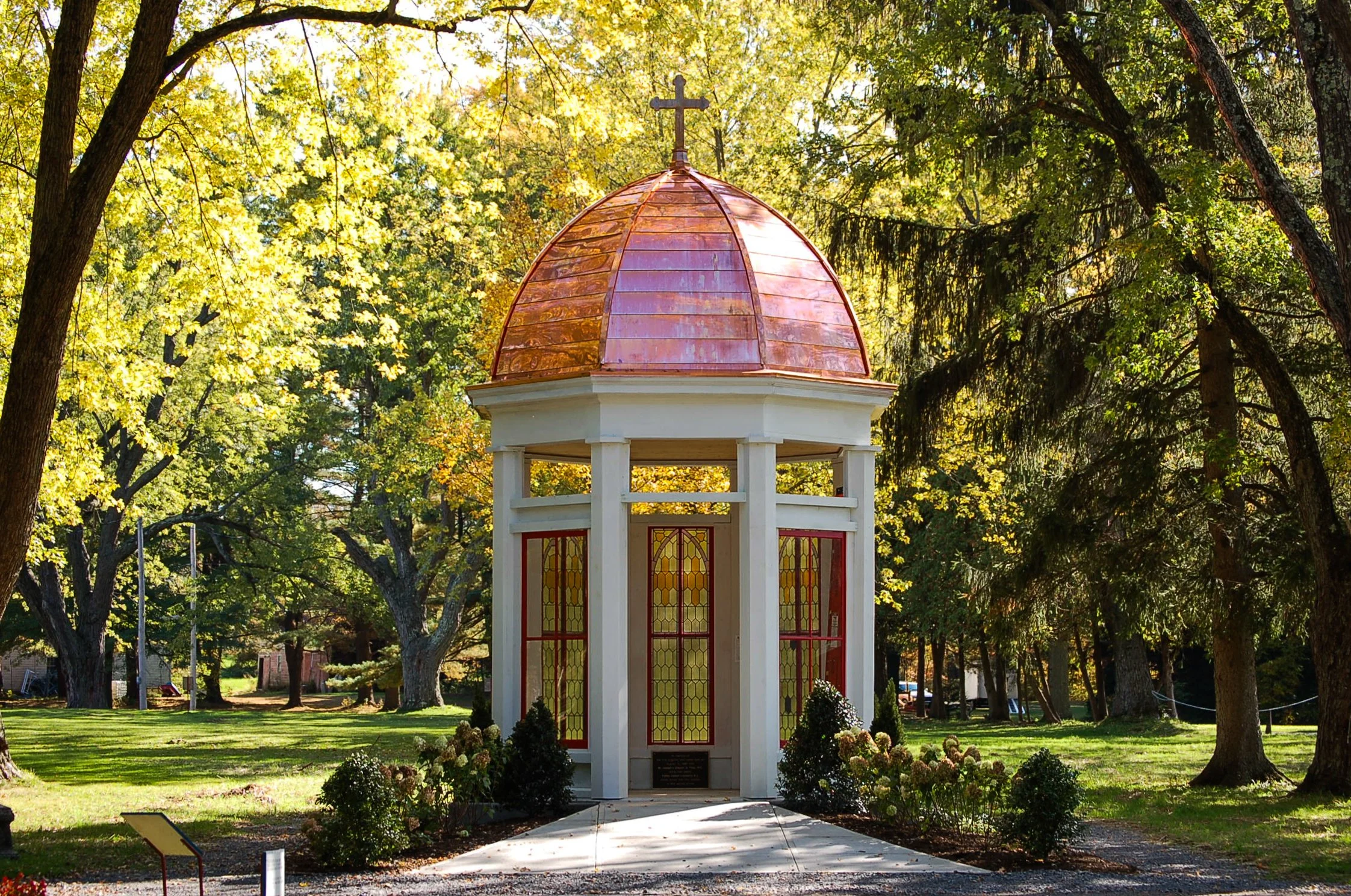 A small, open pavilion with stained glass doors and a copper dome roof with a cross on top, surrounded by lush trees in a park setting.