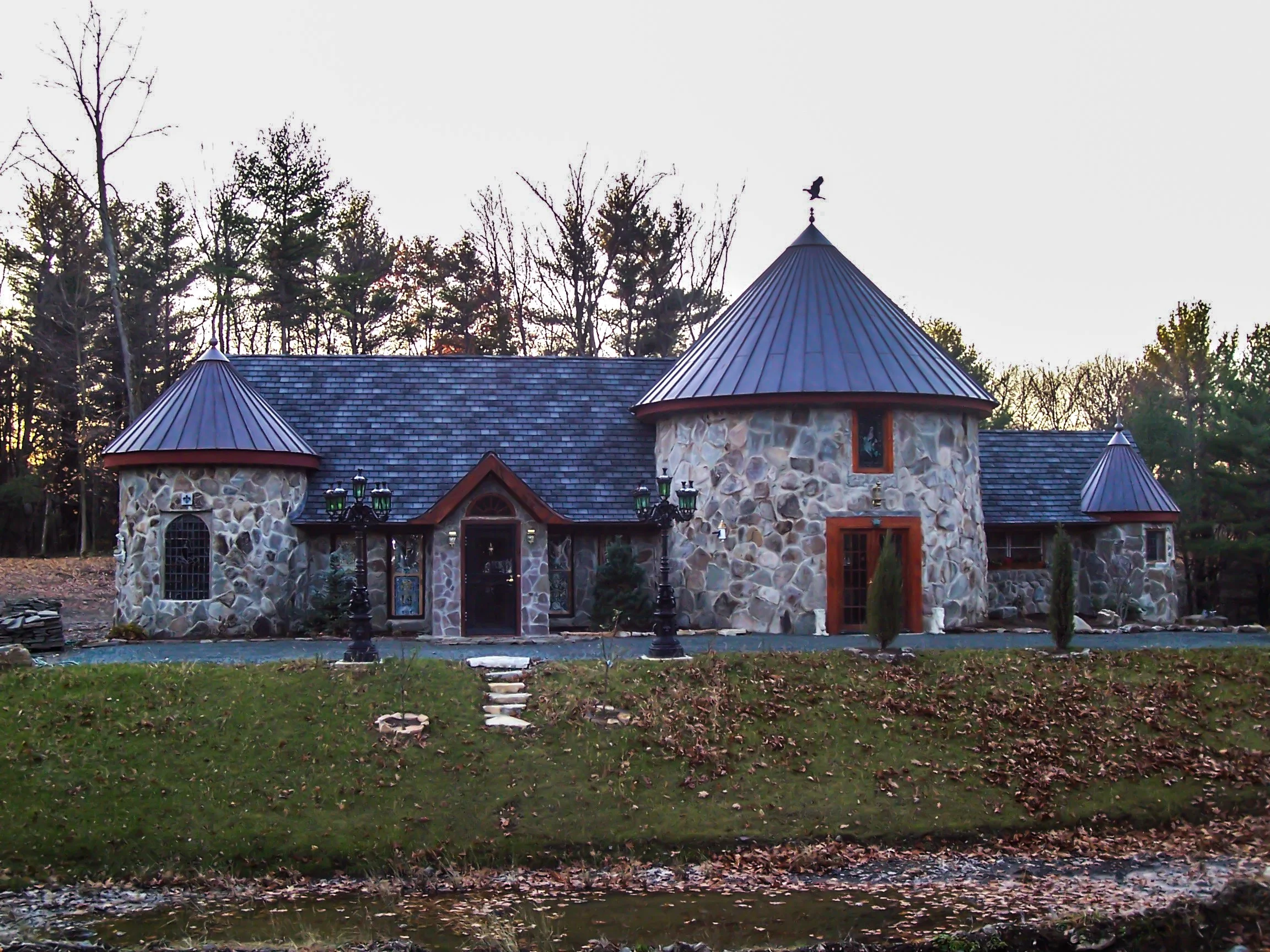 A stone castle-style house with a conical metal roof, small trees in front, and a wooded background.