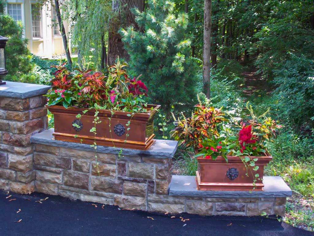 Two large rectangular flower pots with decorative lion head emblems, filled with colorful plants, are placed on a stone wall beside a wooded area.