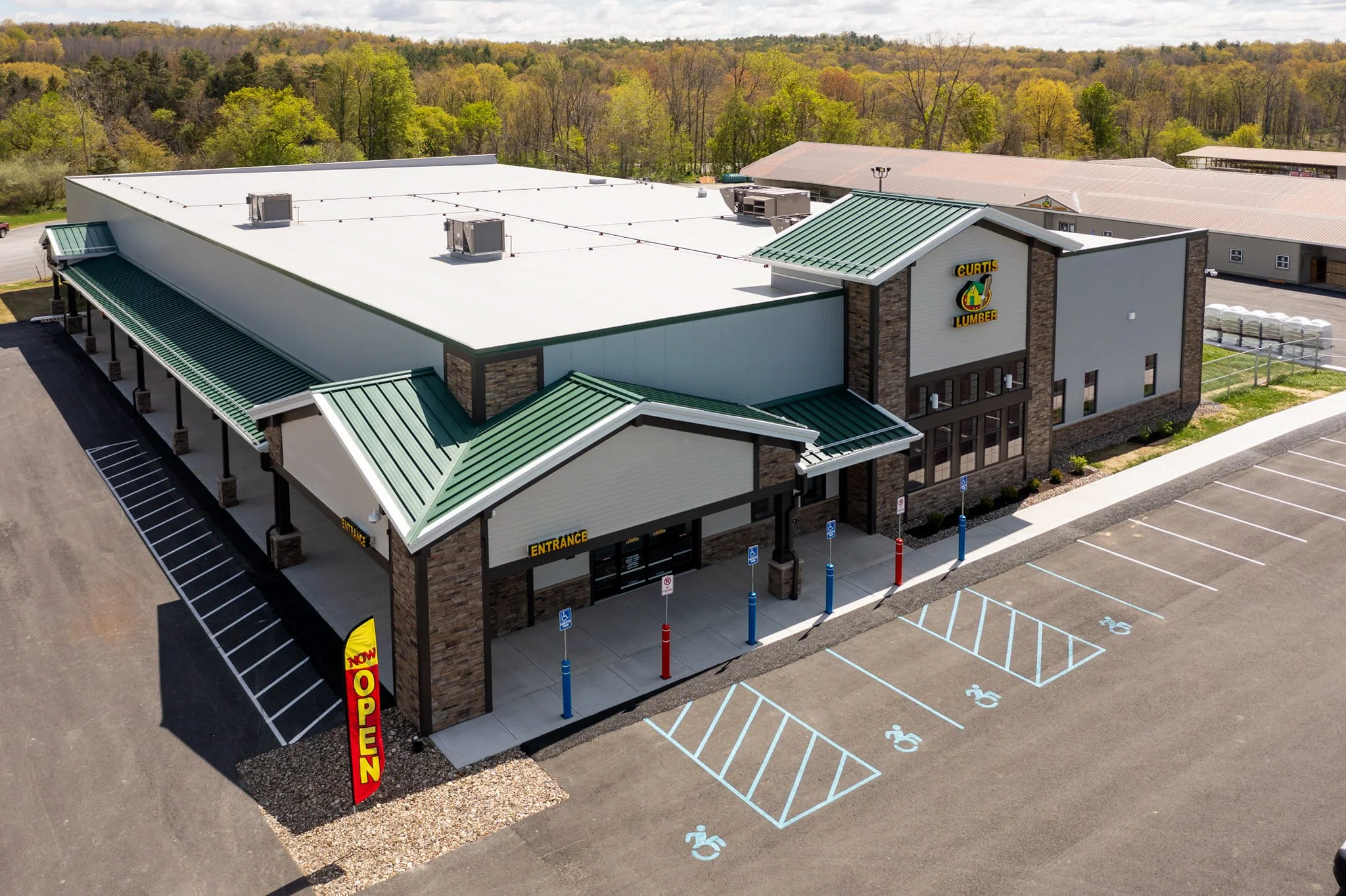 A modern retail store with a green metal roof, brick accents, and an entrance marked with a yellow 'OPEN' flag. The parking lot has designated handicapped spaces and no vehicles present. The store's sign reads 'Curtis Lumber'.