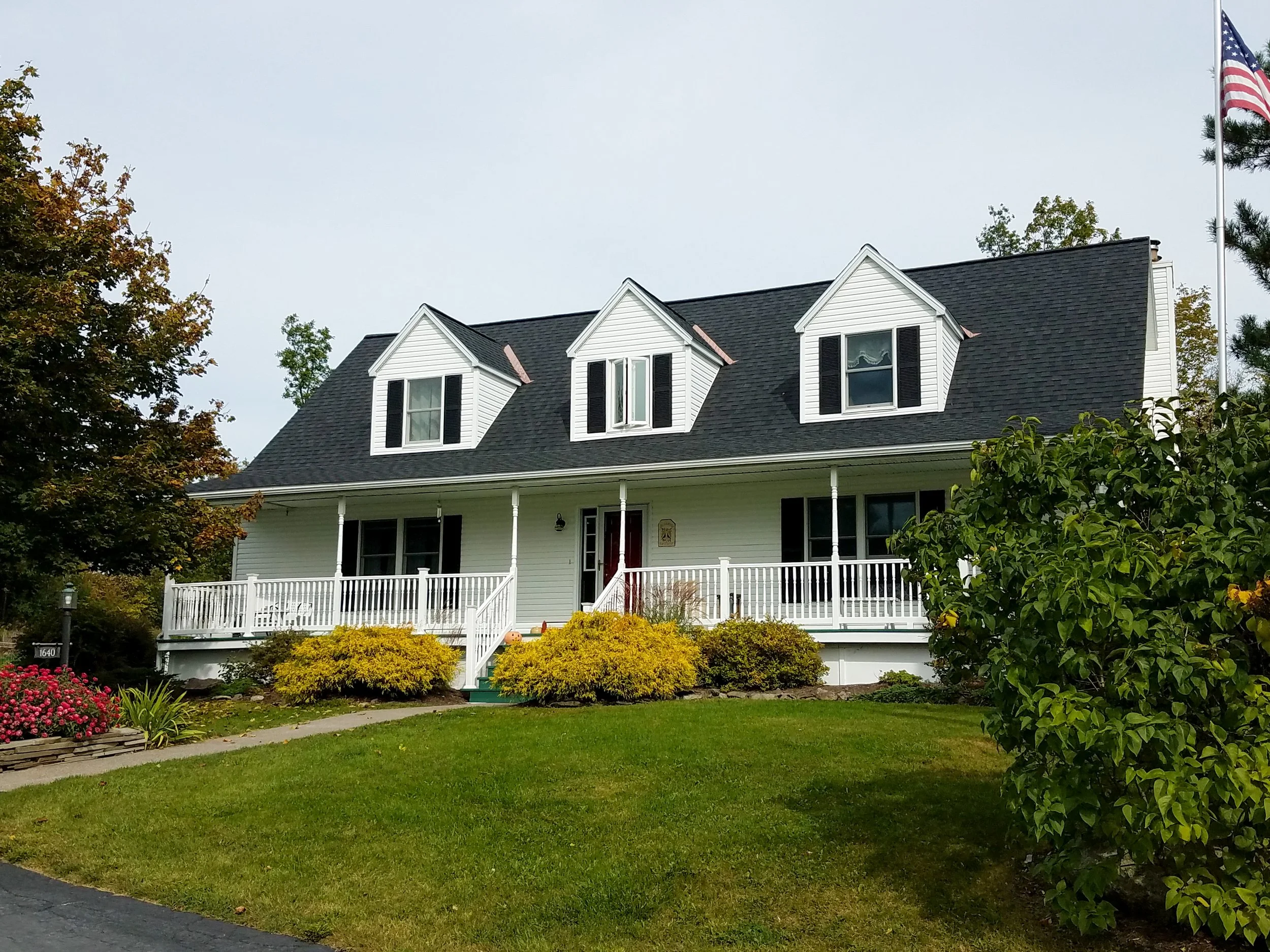 White house with black roof, black shutters, and a porch with white railing. There are green and yellow bushes in front, along with a neighbor's flower bed with pink flowers. A flagpole with the American flag is on the right side of the house.