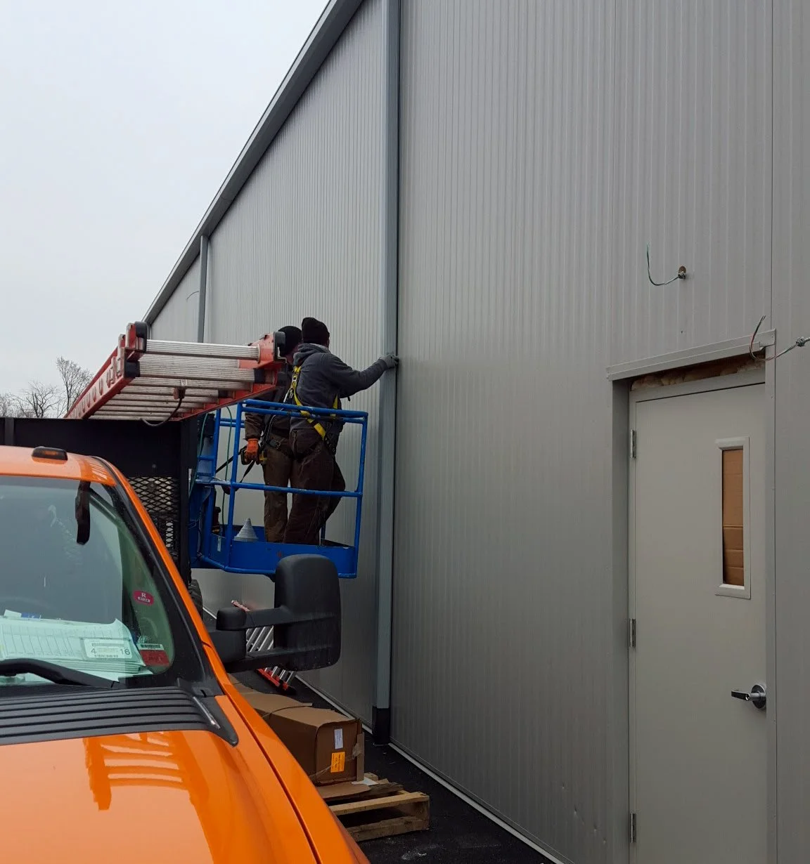 Two workers in safety gear installing or inspecting the exterior wall of a building using a lift.