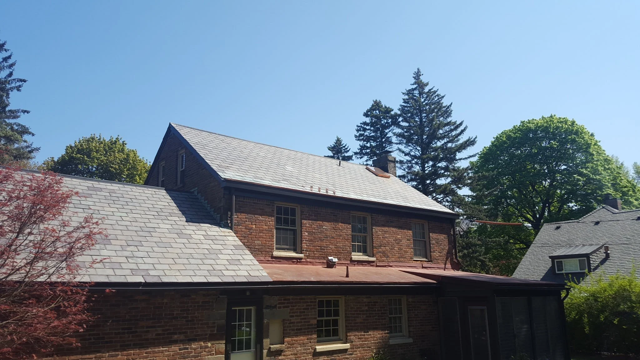 Two-story brick house with gray shingled roof and three windows on the second floor, surrounded by trees and clear blue sky.