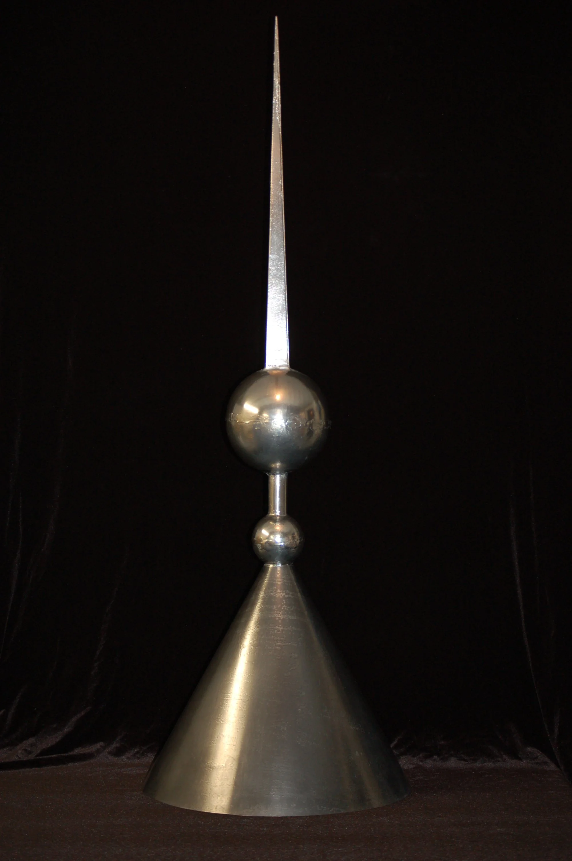 A metallic sculpture consisting of a conical base, two spherical shapes, and a pointed rod on top, set against a dark background.