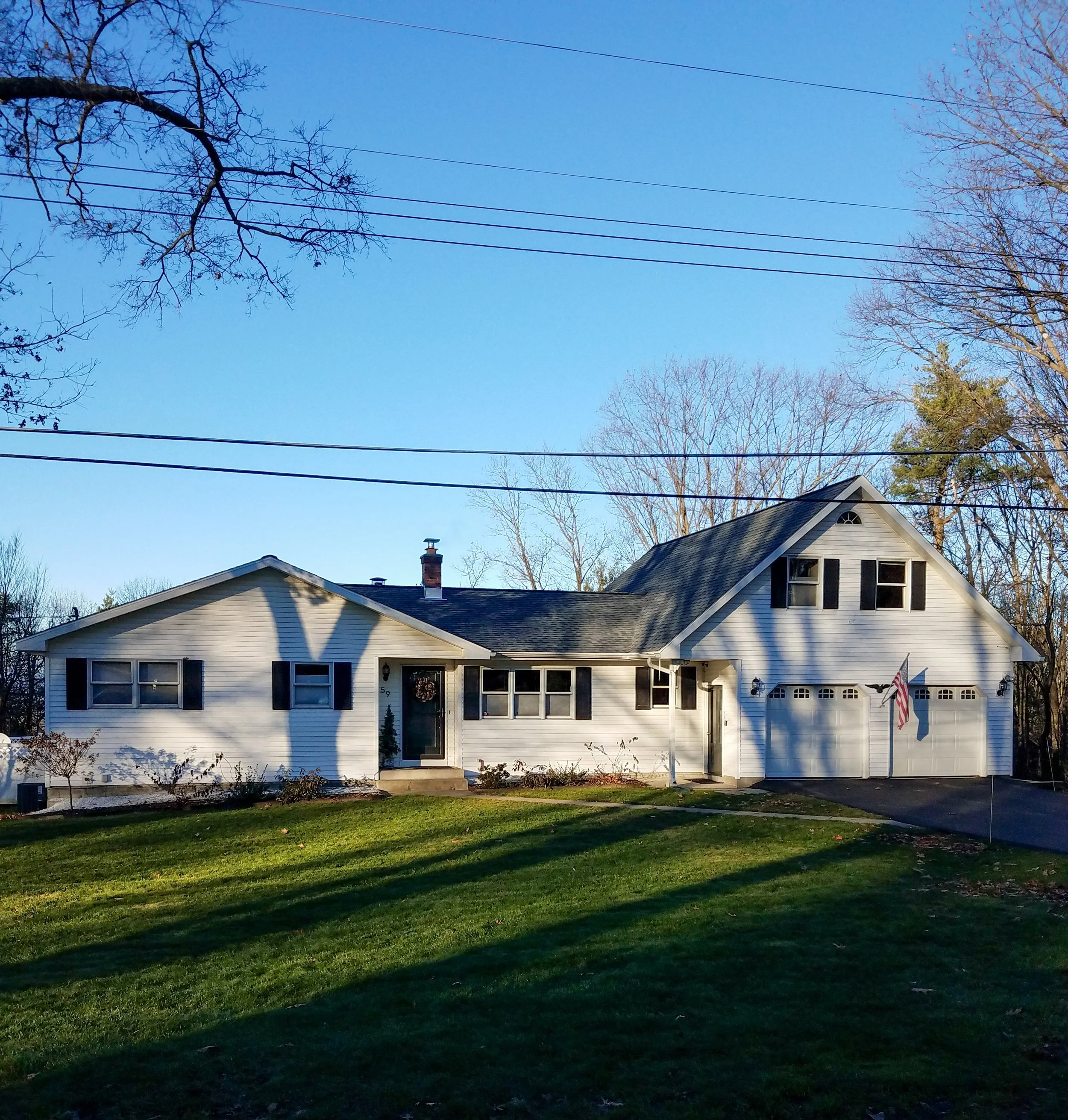 A white suburban house with black shutters, a two-car garage, and a front yard with green grass, in a neighborhood with leafless trees and a clear blue sky.