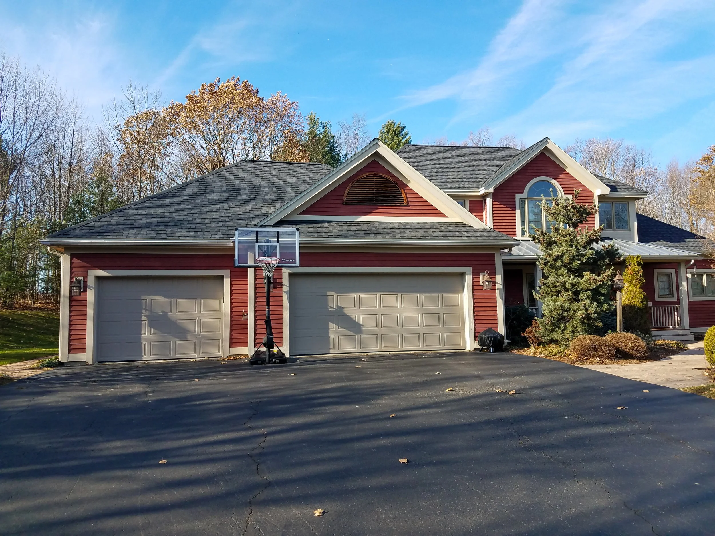 A large house with red siding, two garage doors, and a basketball hoop on the driveway, surrounded by trees with fall foliage.