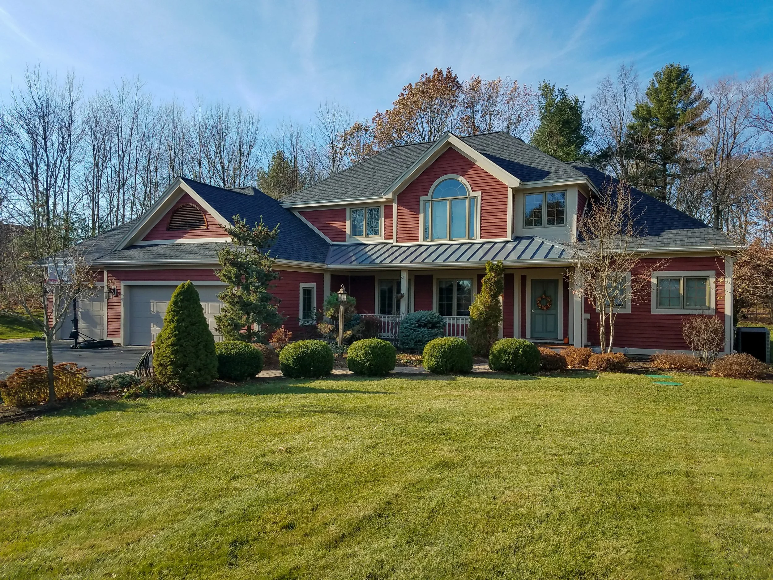 Front view of a two-story suburban house with red siding, gray roof, and white trim, surrounded by a lush green lawn and neatly trimmed bushes, trees, and garden