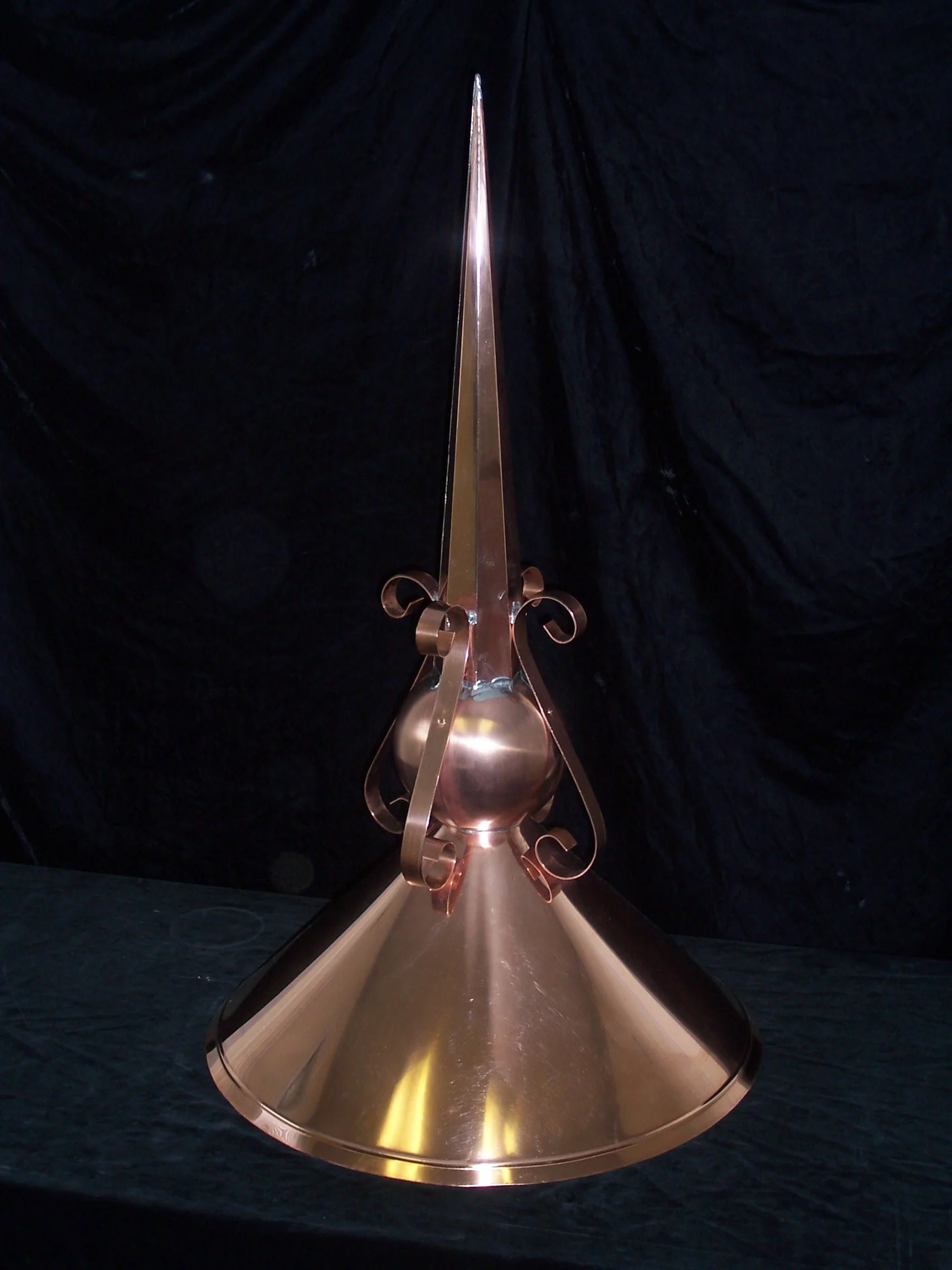 A metallic, pointed ceremonial object with a conical base and a tall, slender spire, set against a dark background.