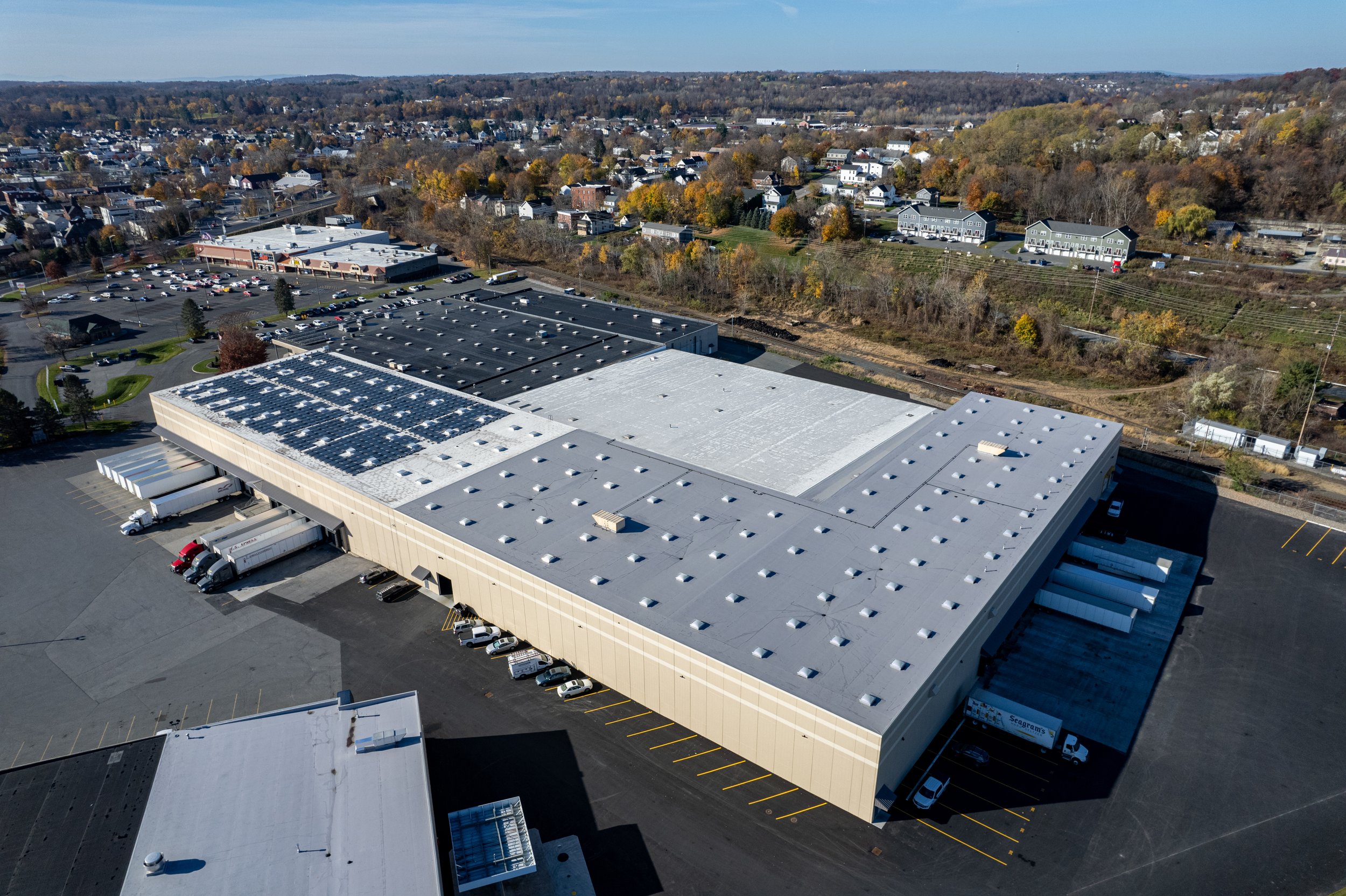 An aerial view of a large industrial warehouse with solar panels on part of its roof, surrounded by parking lots with several trucks and cars, and a landscape of a suburban area with houses and trees in the background during daytime.