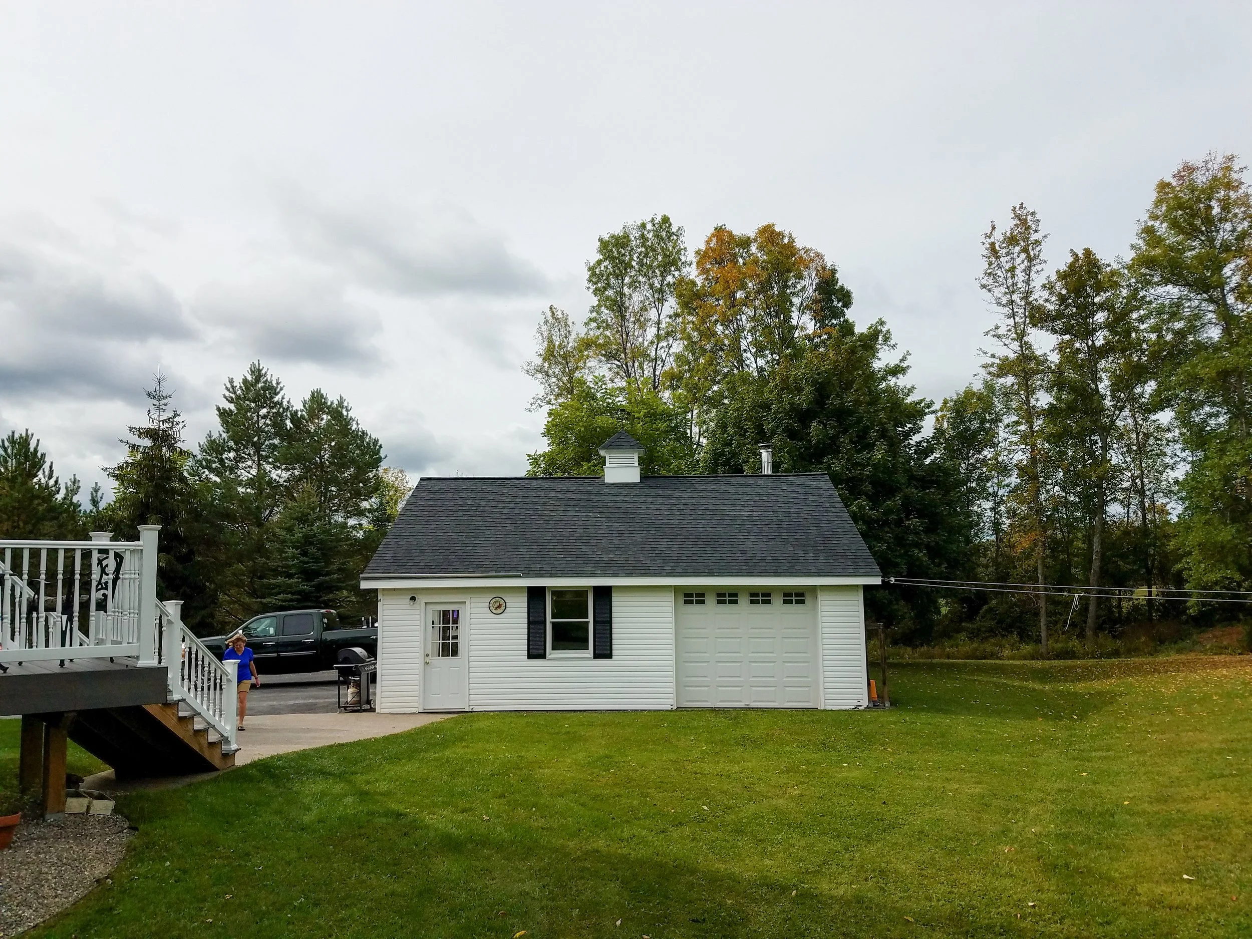 White house with black roof, garage door, and a woman standing outside in a grassy yard with trees in the background.