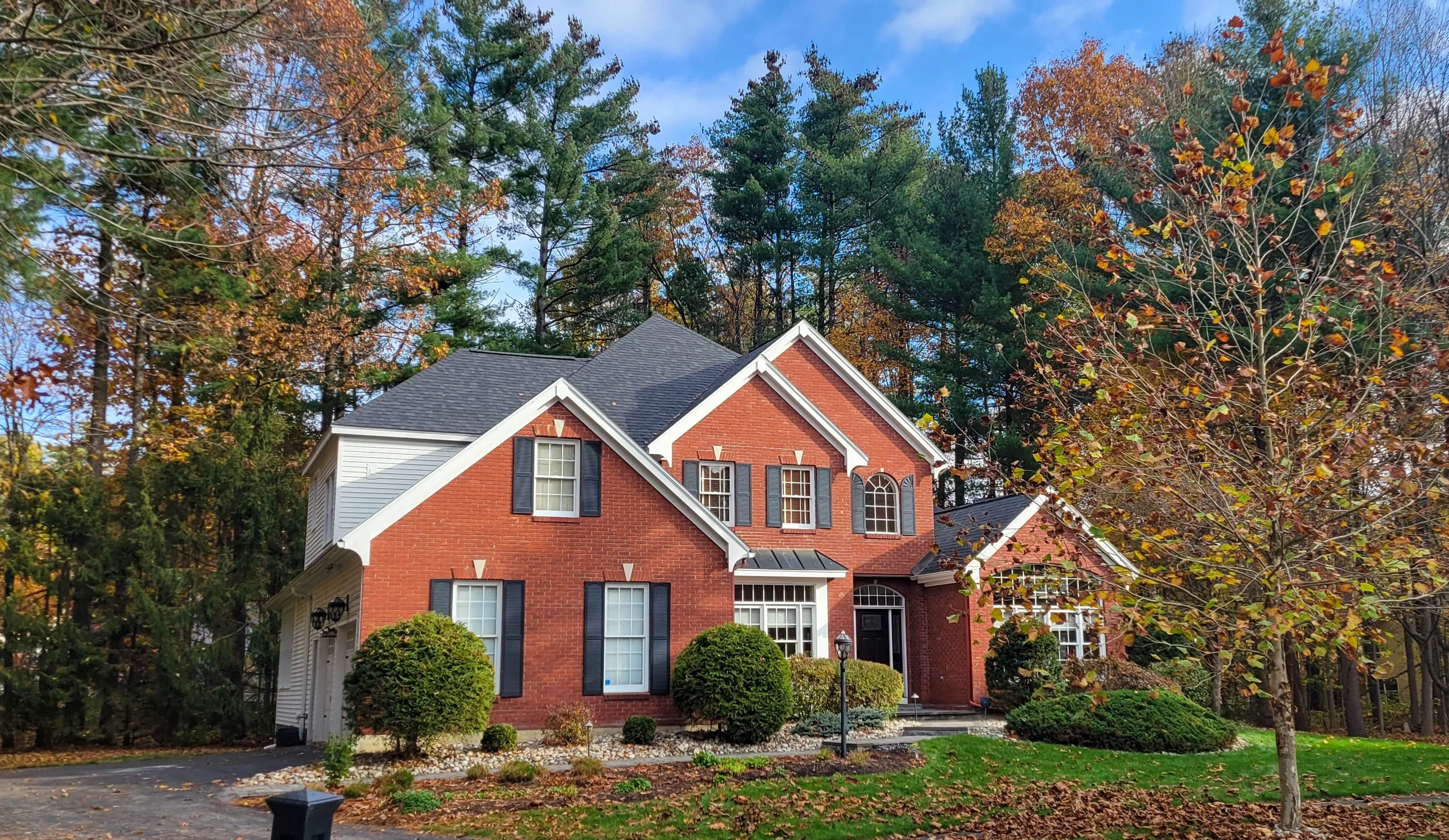 A two-story brick house with white trim, black shutters, surrounded by trees with autumn foliage. A small front yard with bushes, a lamp post, and a stone walkway leading to the entrance.
