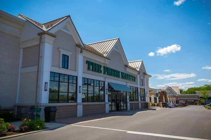 Exterior of a grocery store called The Fresh Market on a sunny day, with parking lot and cars in front.