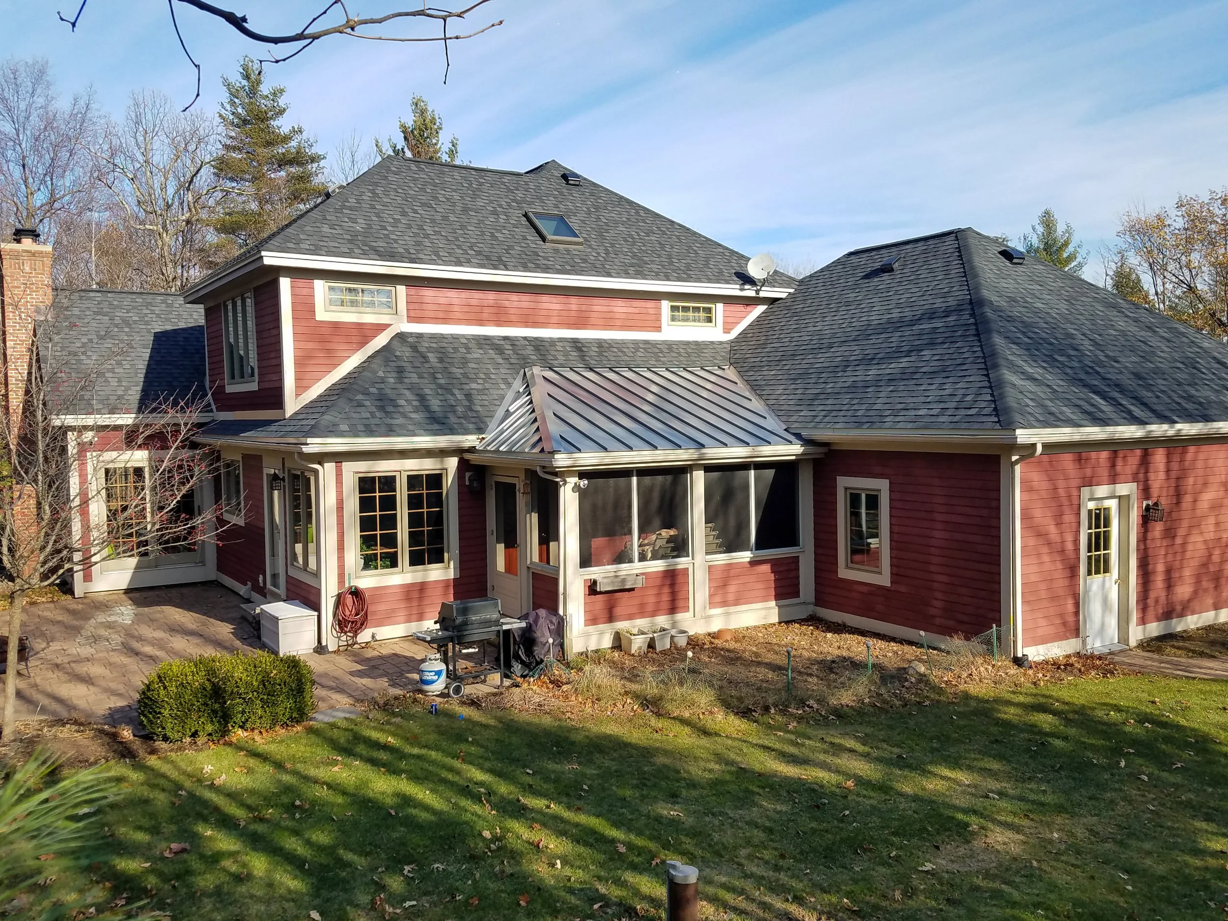 Back of a two-story house with red siding, multiple windows, and a sloped roof. The house has a screened porch with a metal roof and a small paved patio area with gardening supplies. There is grass, a small bush, and some trees with autumn leaves in 