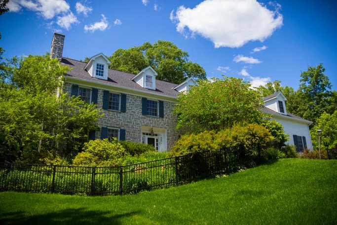 A large house with stone and white siding, surrounded by green trees and bushes, under a bright blue sky with a few clouds, and enclosed by a black metal fence.