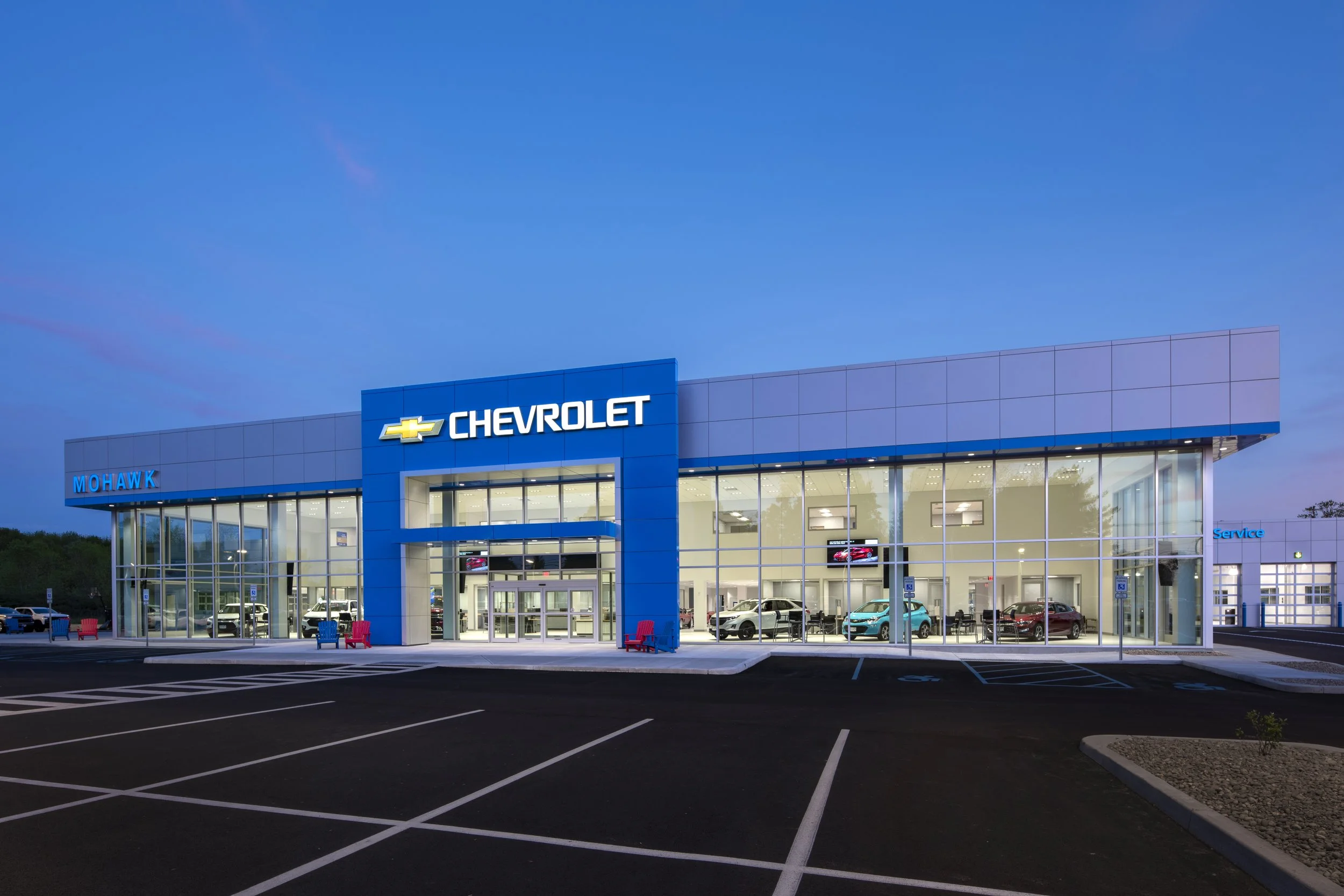 Exterior view of a Chevrolet car dealership building during dusk, with a large Chevrolet sign and glass windows showcasing cars inside, and an empty parking lot in the foreground.