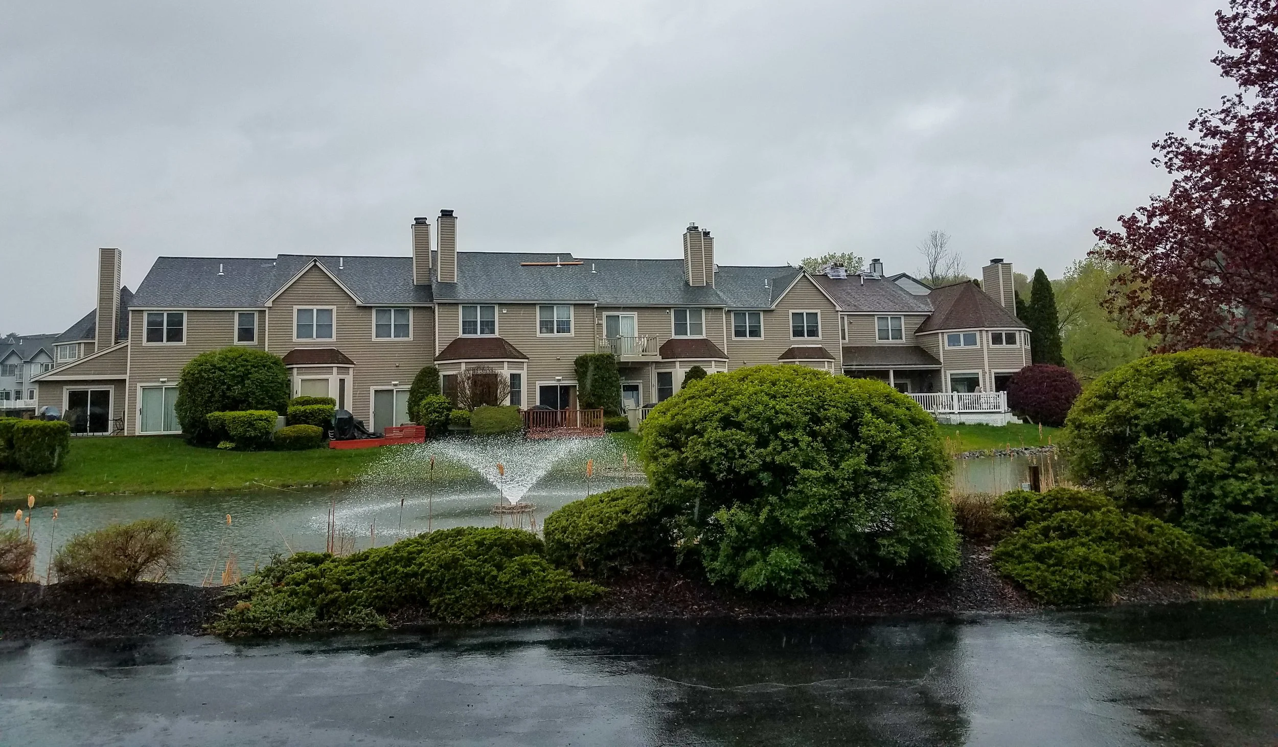 Multi-story beige residential building with multiple chimneys, surrounded by well-maintained bushes and trees, next to a small pond with a fountain, on a rainy day.