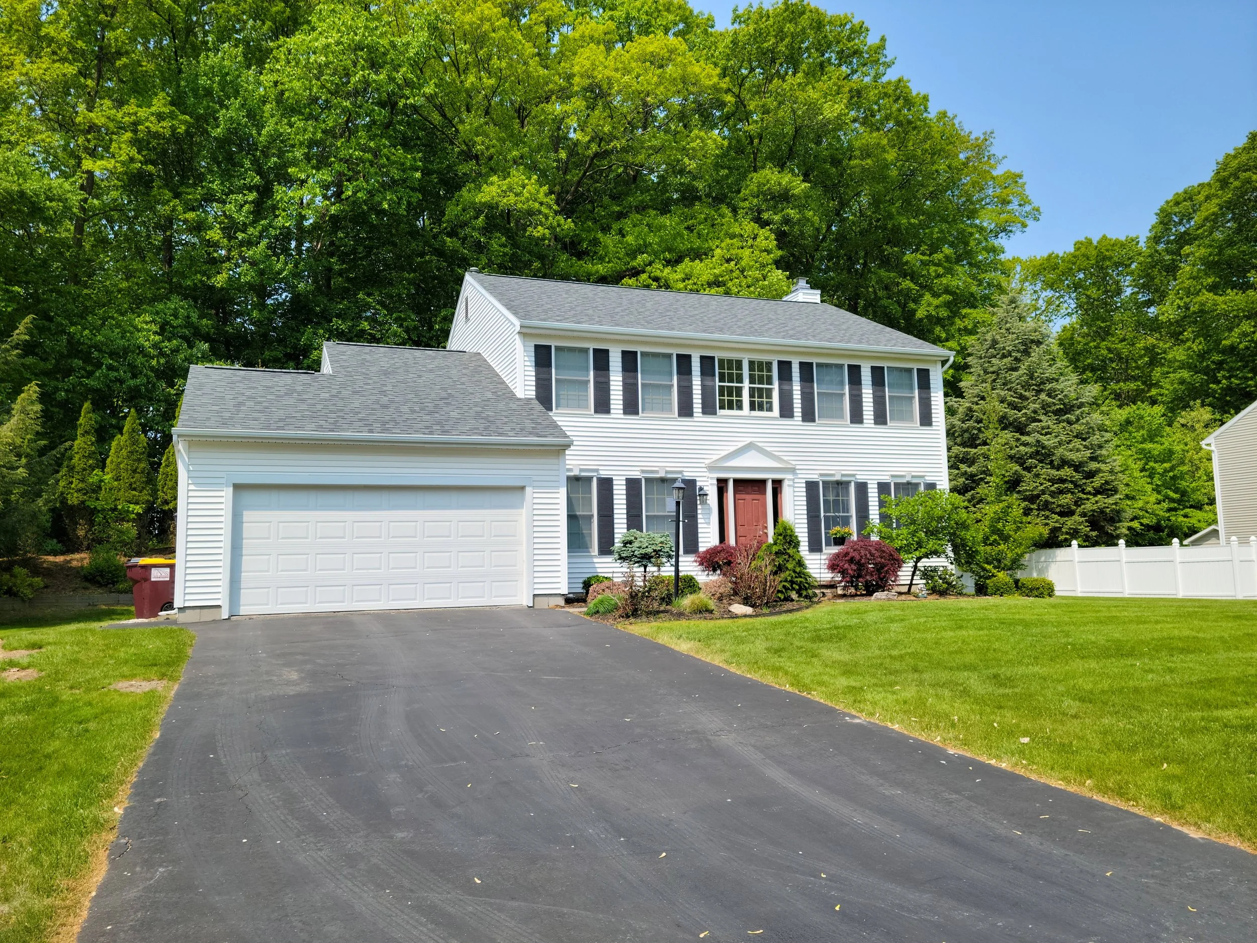 White two-story house with attached garage, surrounded by green lawn and trees, on a bright sunny day.