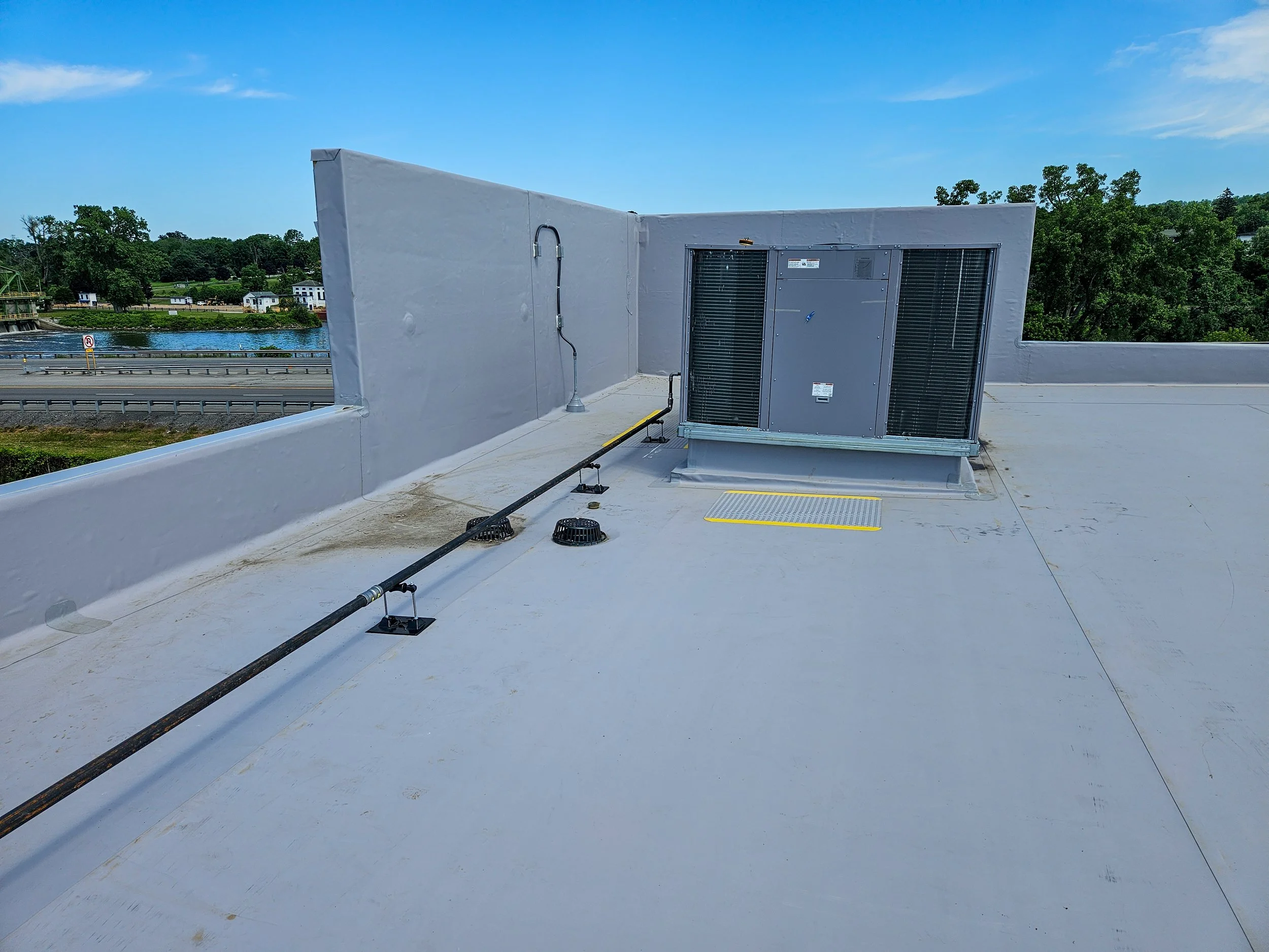 Rooftop with HVAC unit, cables, and drains, overlooking a highway, river, and trees on a blue sky day.