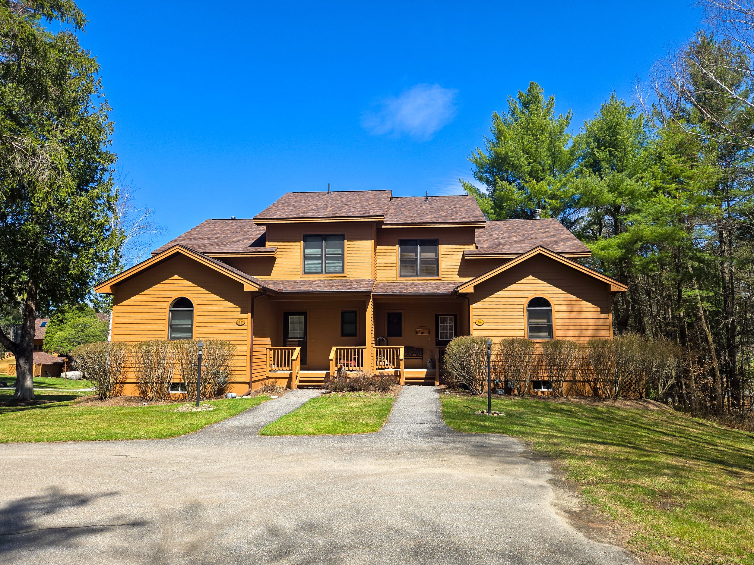 A large, two-story house with yellow siding and brown shingled roof, surrounded by trees and bushes, with a driveway leading up to the front porch under a bright blue sky.