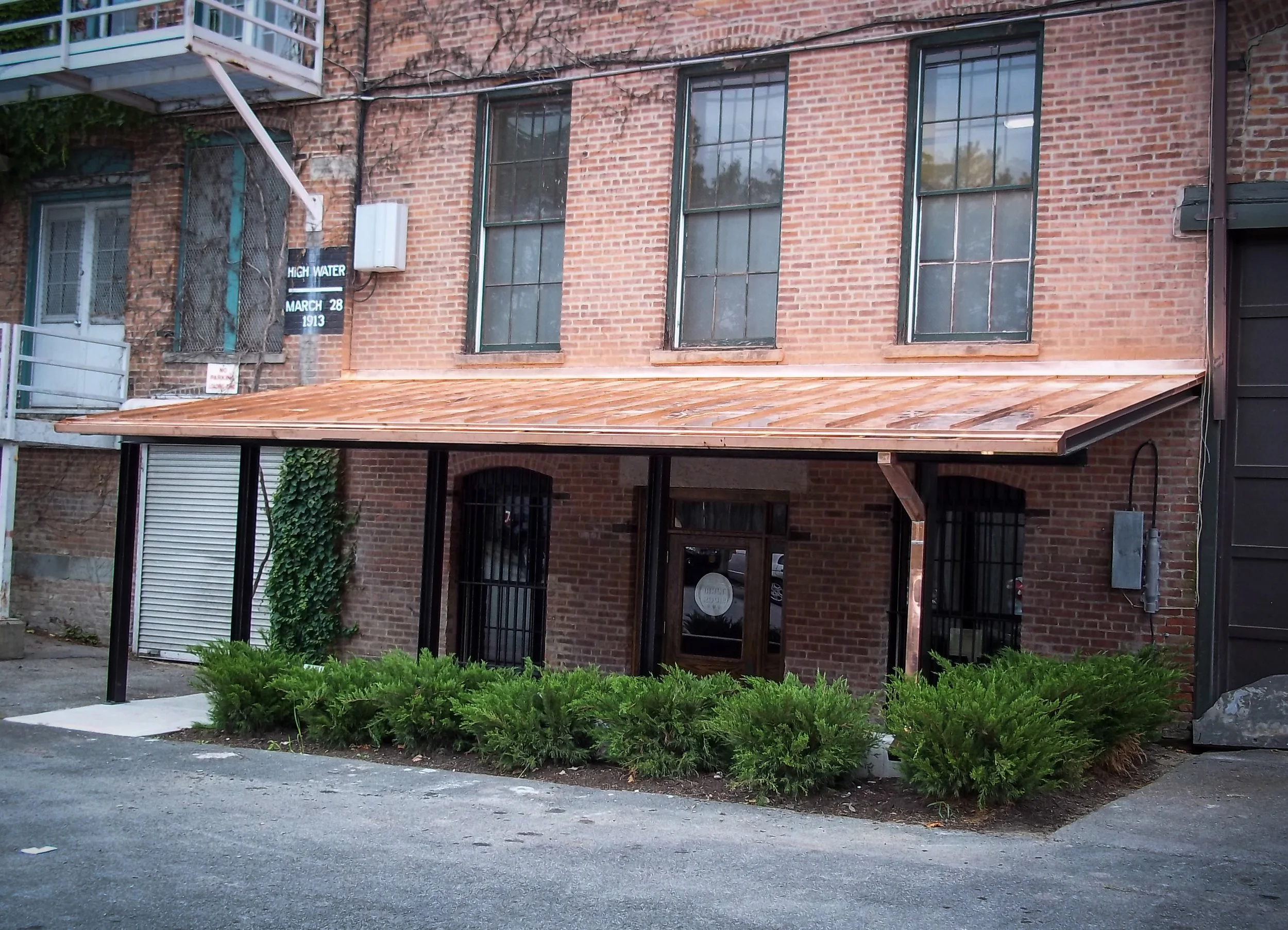 Brick building with large windows and a wooden awning above the entrance, surrounded by green bushes in front.