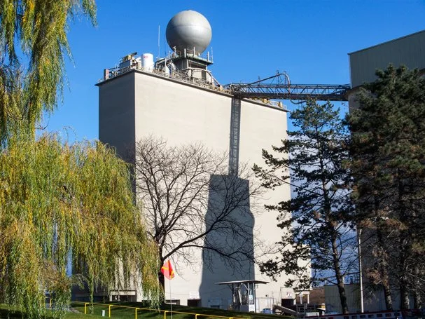 A large building with weather radar equipment on top, surrounded by trees and a partly cloudy blue sky.