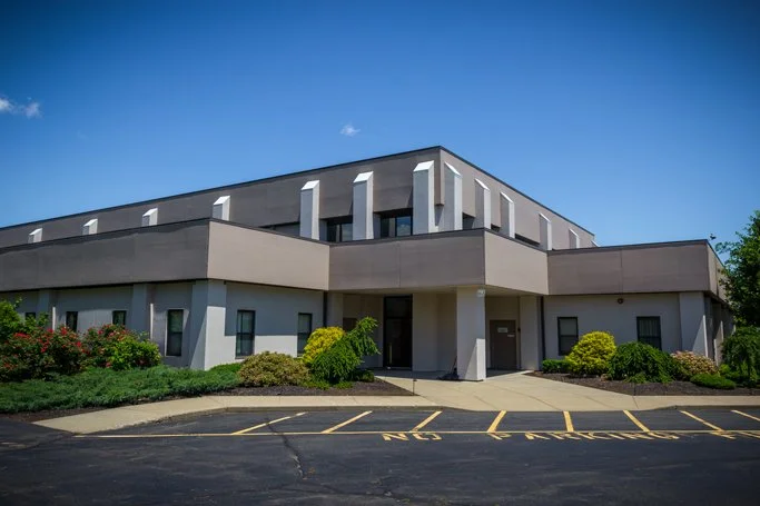 Front view of a modern, multi-story office building with landscaped bushes and a parking lot in front.