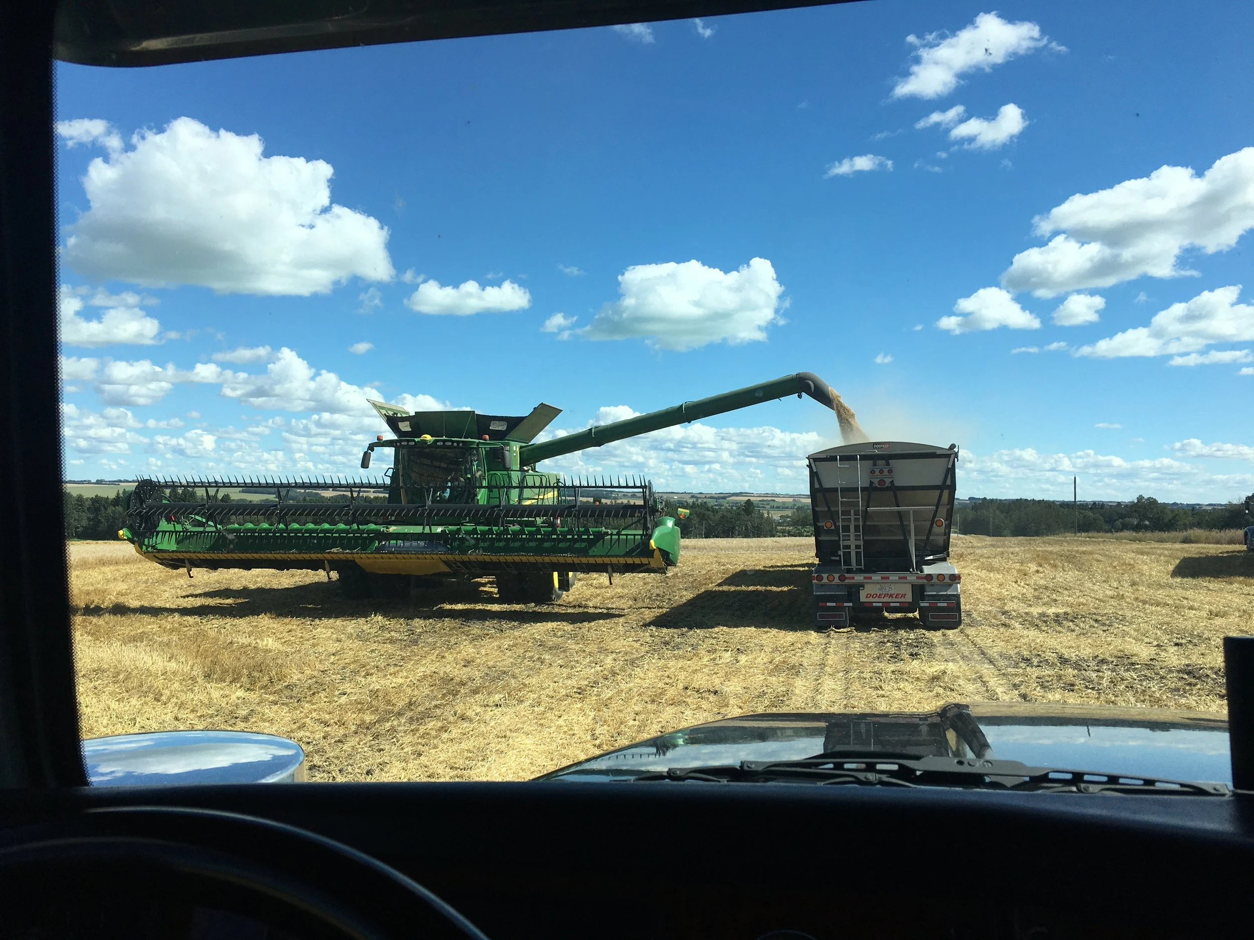 John Deere combine harvester unloading grain into grain truck on a farm field under a blue sky with clouds.