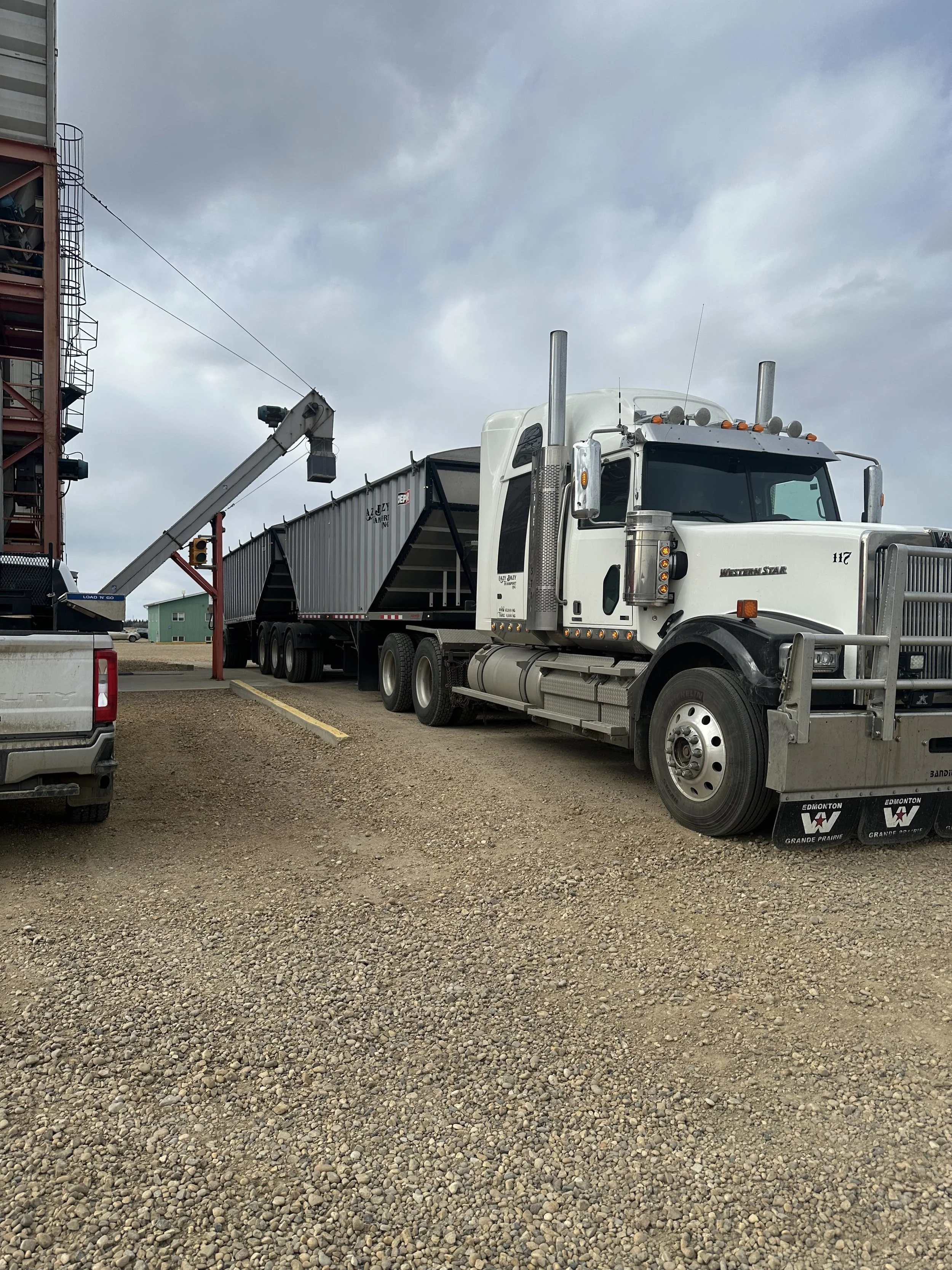 A large white semi-truck with a gray trailer parked on gravel next to a building with a tall tower. The truck has a boat or equipment loads on the trailer, and there is a crane arm extending from the building toward the trailer.