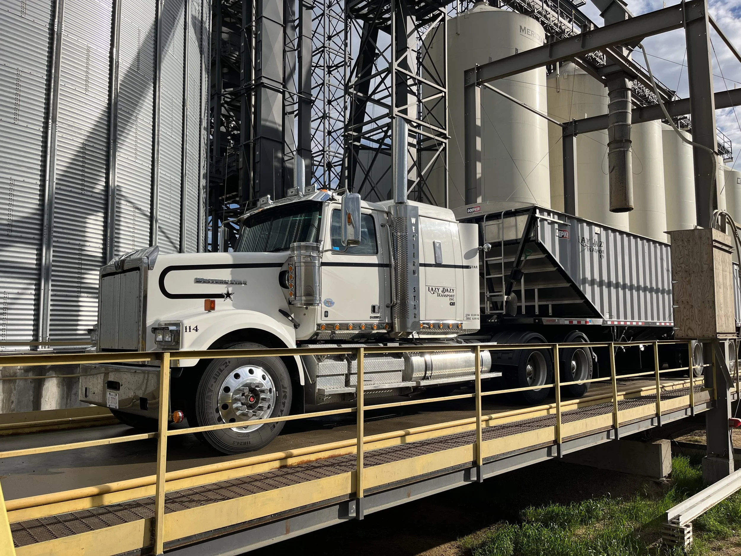 A white semi-truck with a black stripe and a cargo trailer attached, parked on a yellow metal platform in front of industrial silos and metal structures.