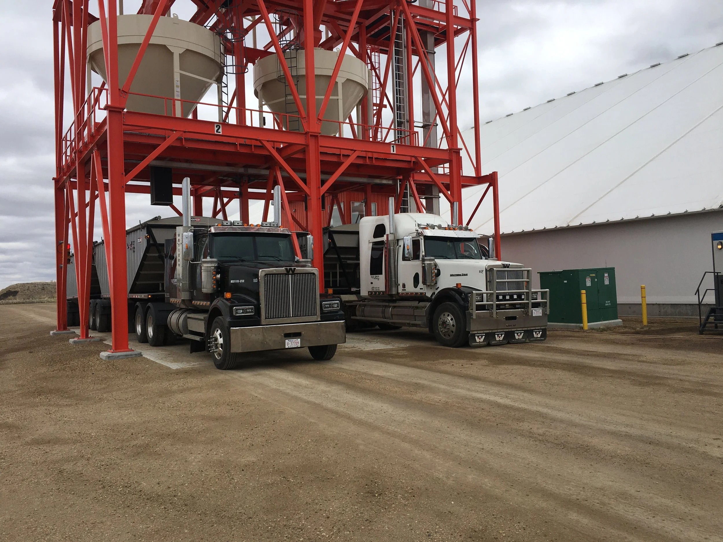 Two large trucks, one black and one white, are parked under a tall red grain silo structure on a gravel lot.