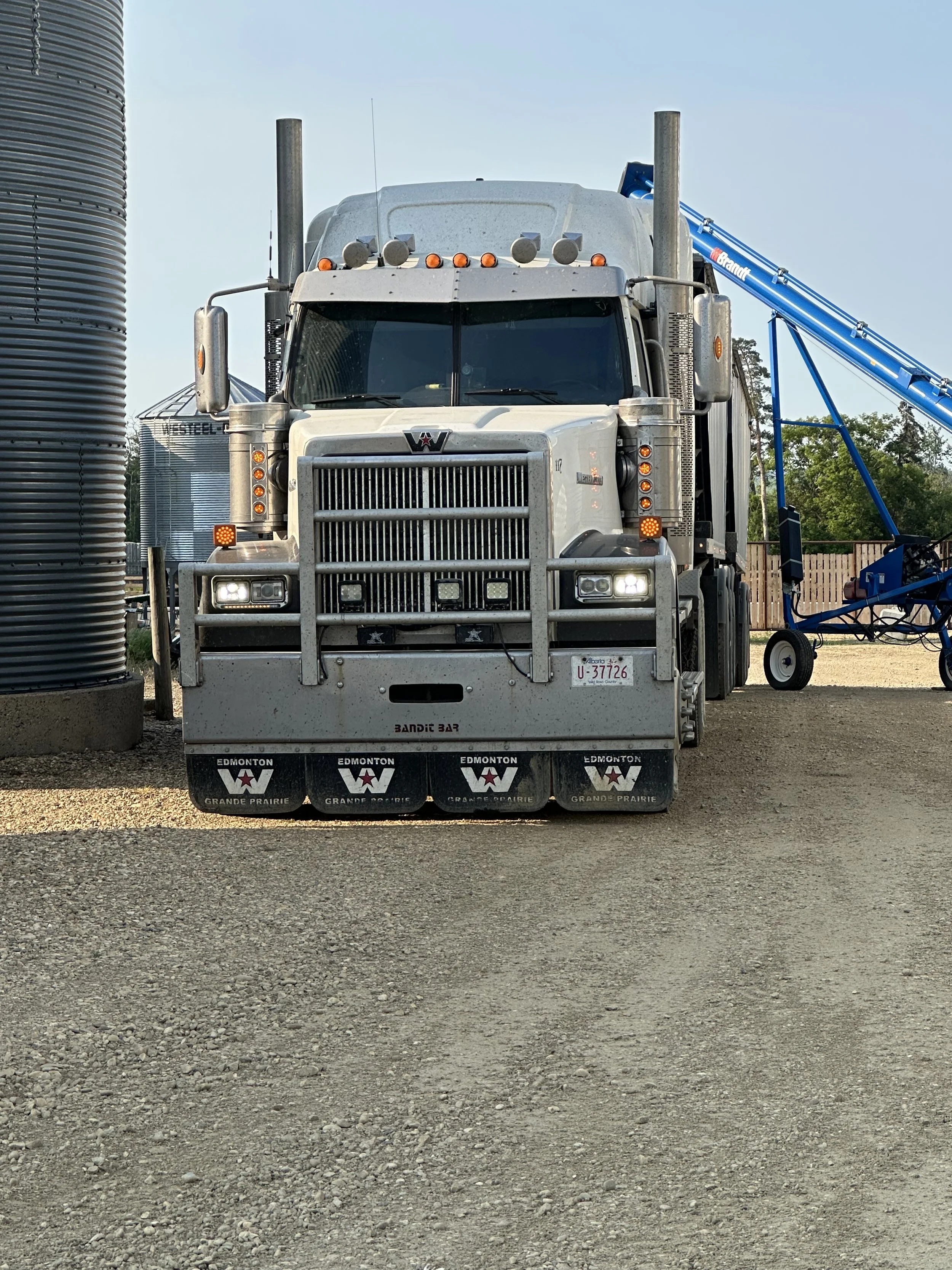 Front view of a large white semi-truck parked outdoors on a gravel surface with a grain silo on the left and farming equipment on the right.