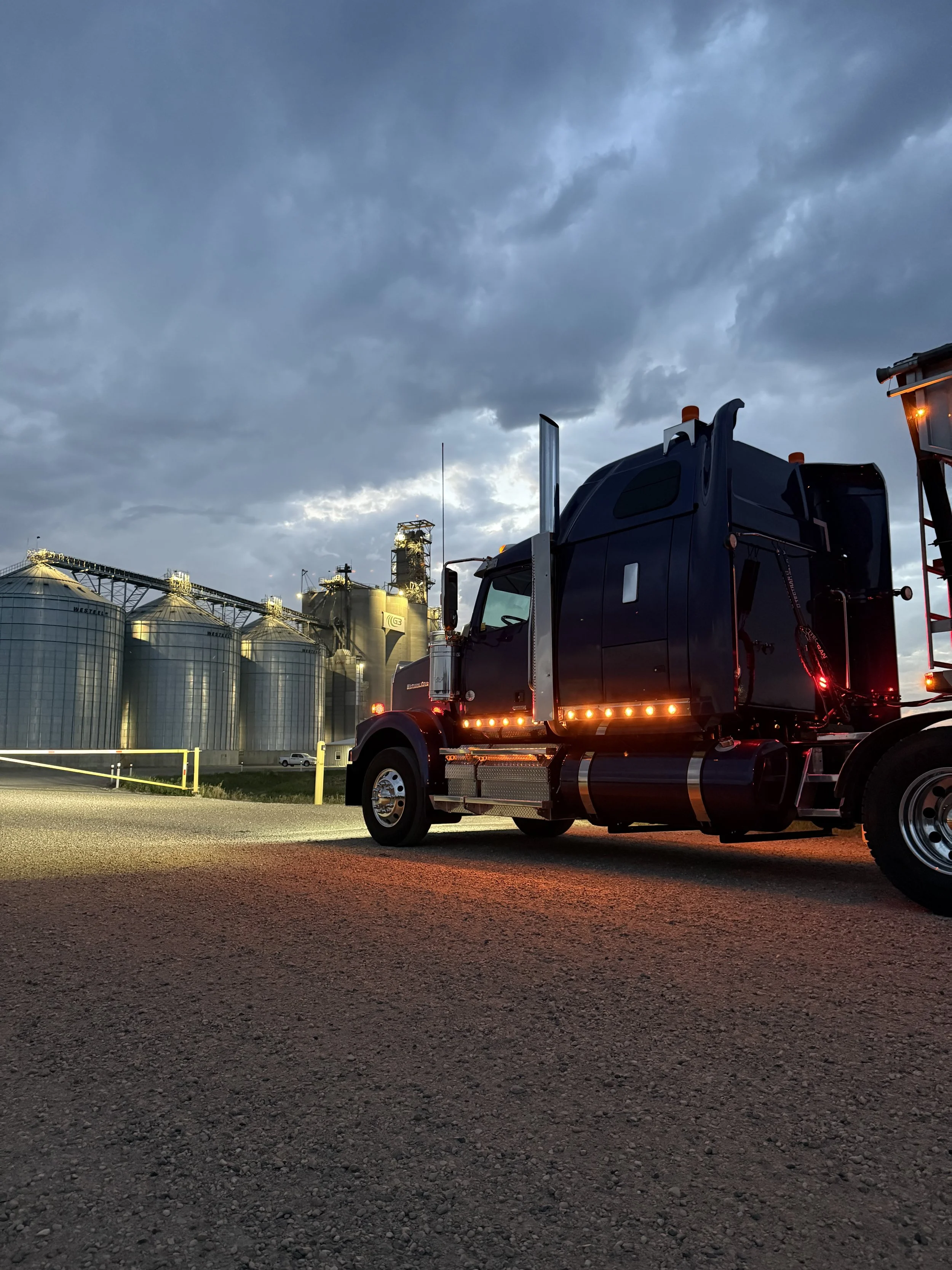 A dark blue semi-truck parked outdoors at dusk with industrial silos and cloudy sky in the background.
