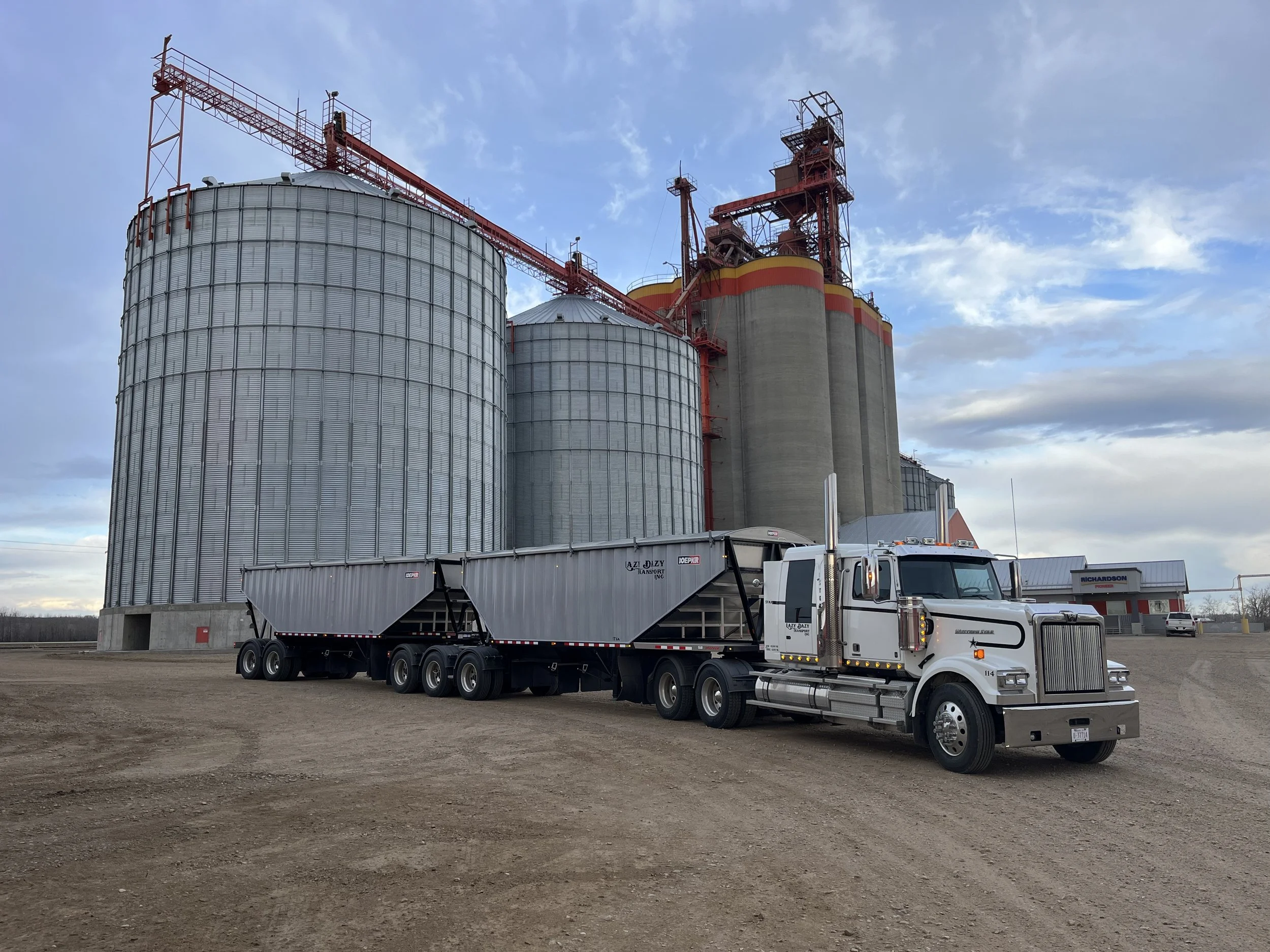 A white semi-truck with a black trailer parked in front of large grain silos on a dirt lot, with a cloudy sky overhead.