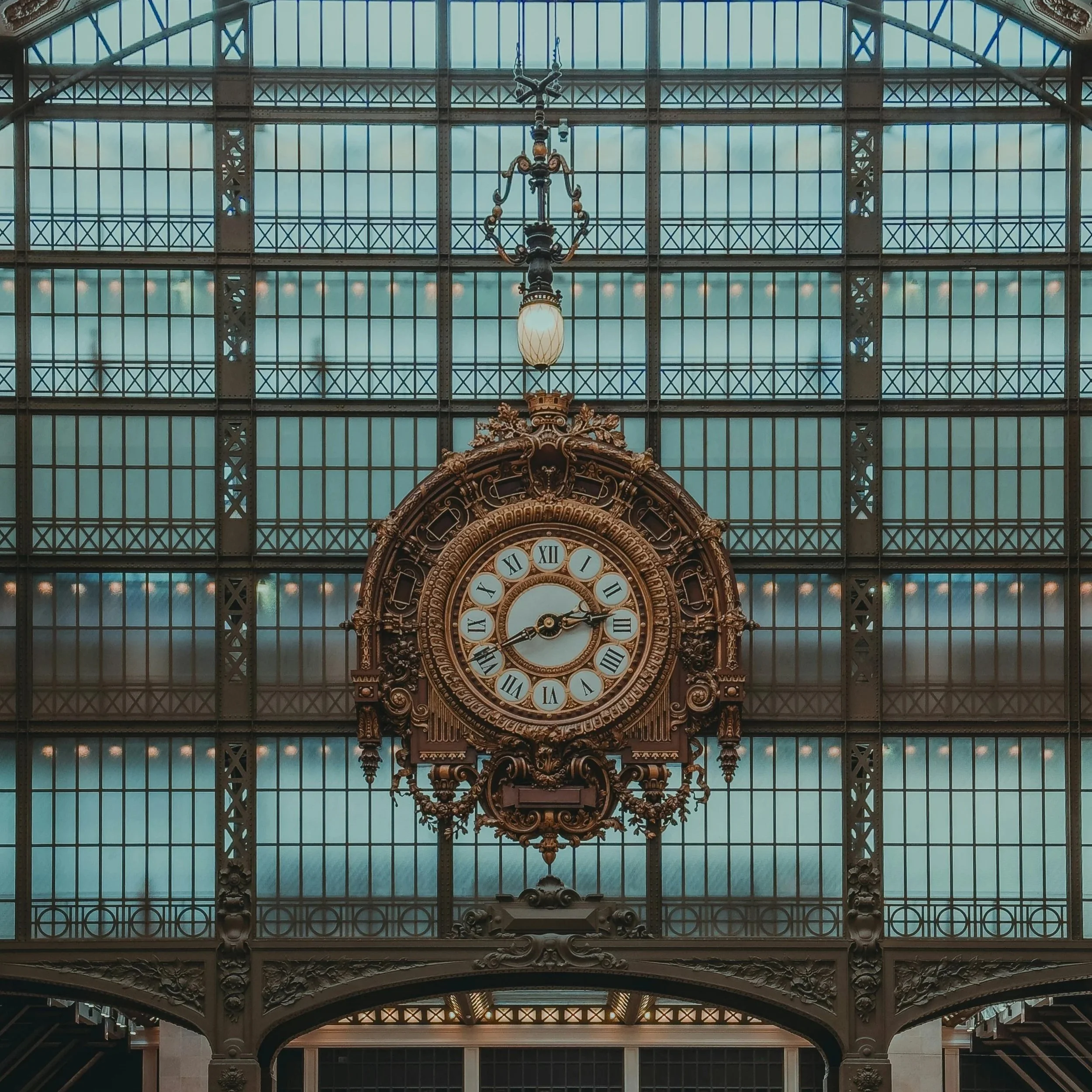 Clock tower surrounded by green leafy trees, with a streetlamp visible on the right side, during daytime.