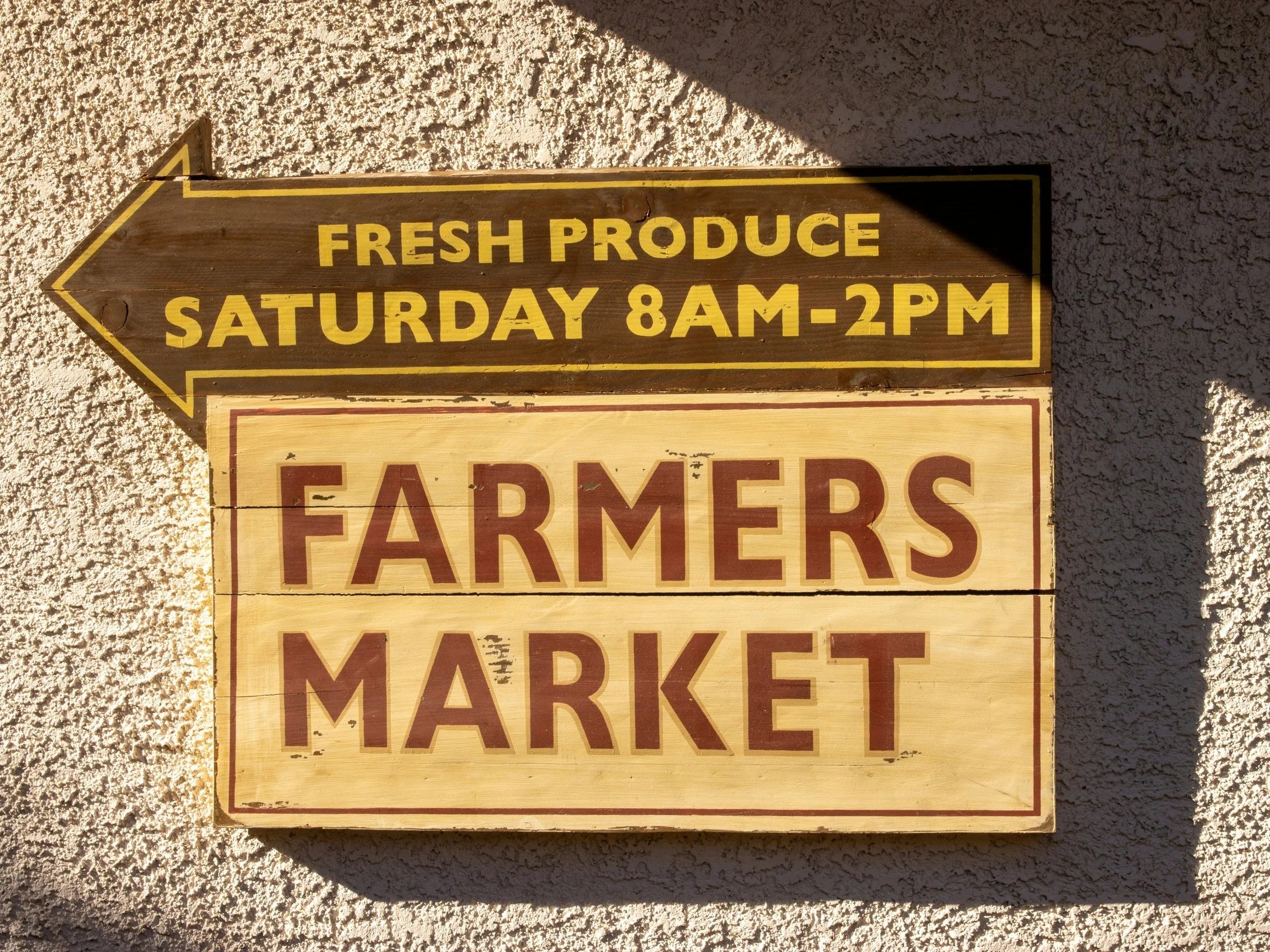 Fresh vegetables including tomatoes, peppers, eggplants, turnips, and radishes displayed on a wooden table in a farm stand with chalkboard signs and green herbs in the background.