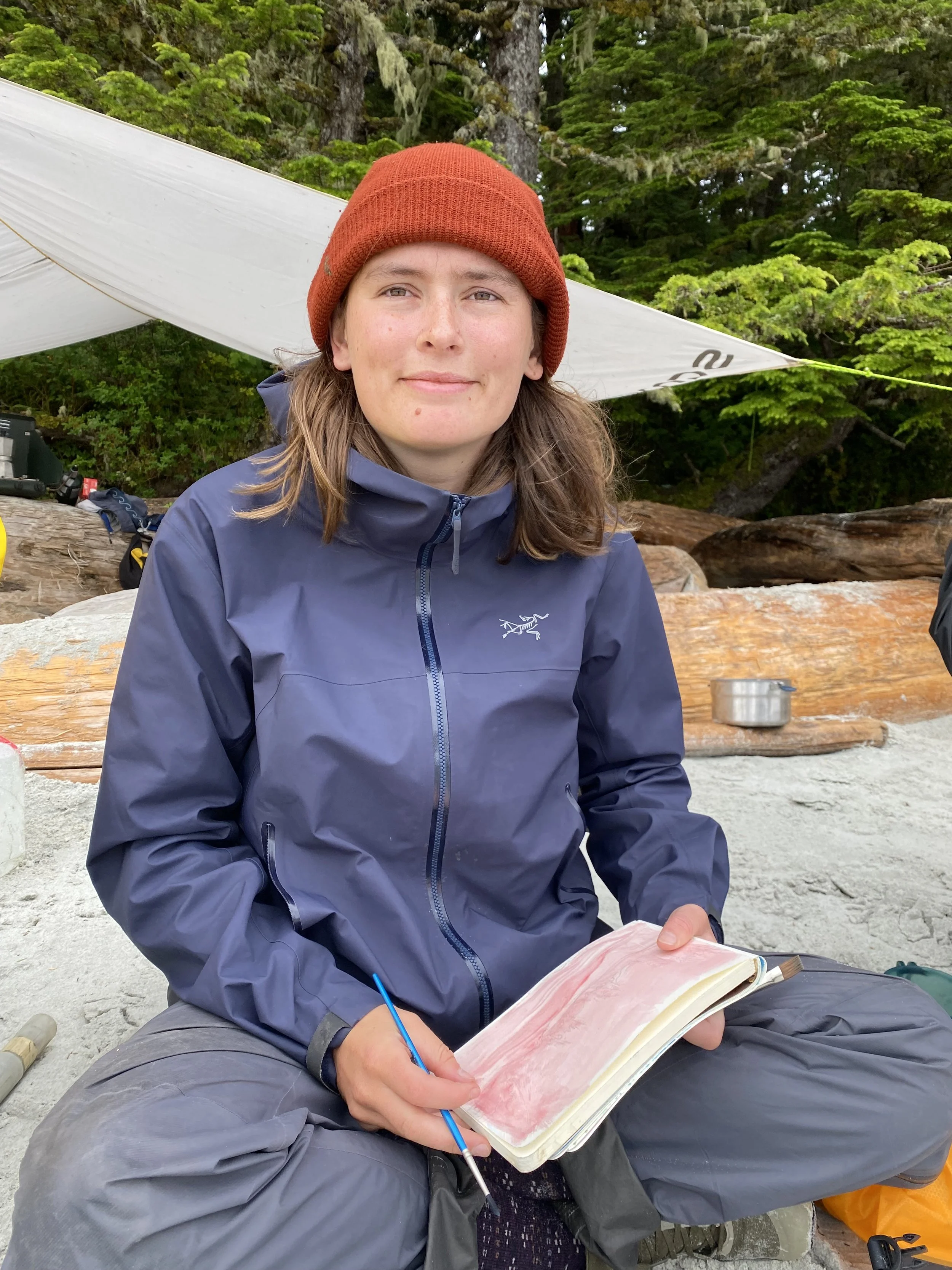 Young person with long brown hair, wearing a red beanie and blue outdoor jacket, sitting on sandy ground surrounded by trees, holding a sketchbook with a pink and white drawing, outdoors in a forested area.