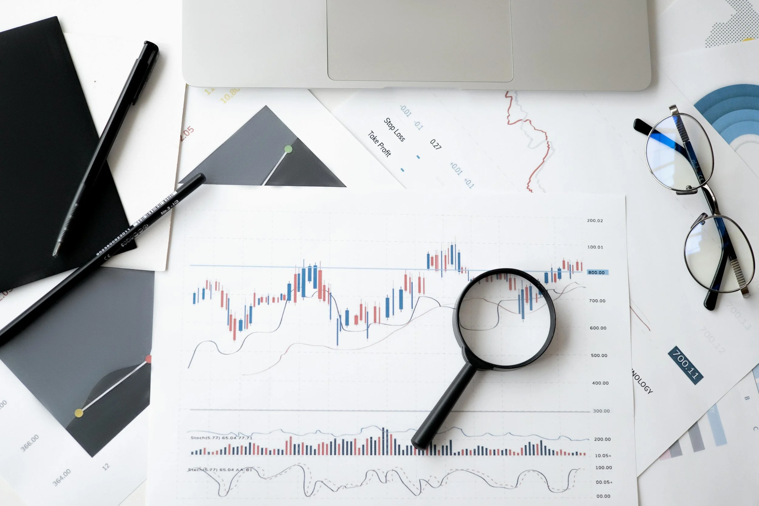 Financial documents with stock charts, a magnifying glass, eyeglasses, pens, and a closed notebook on a white desk.
