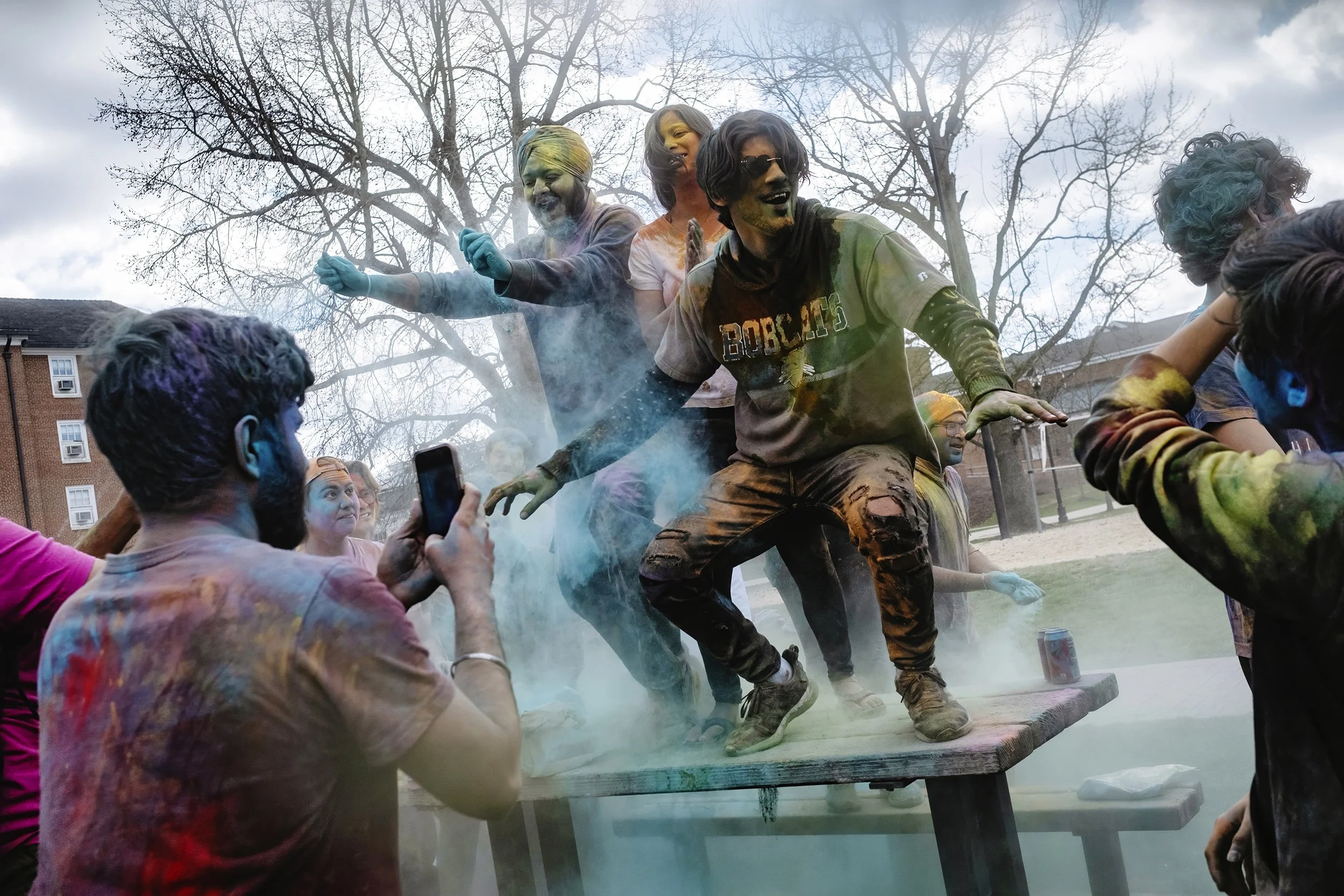 Students Prabhleen Singh Bebi, Sonali Jha, and Harshwardhan Lnu dance on top of a picnic table amid colored powder on the South Green port of Ohio University's campus in Athens, Ohio, on Saturday, March 4, 2023. | Loriene Perera