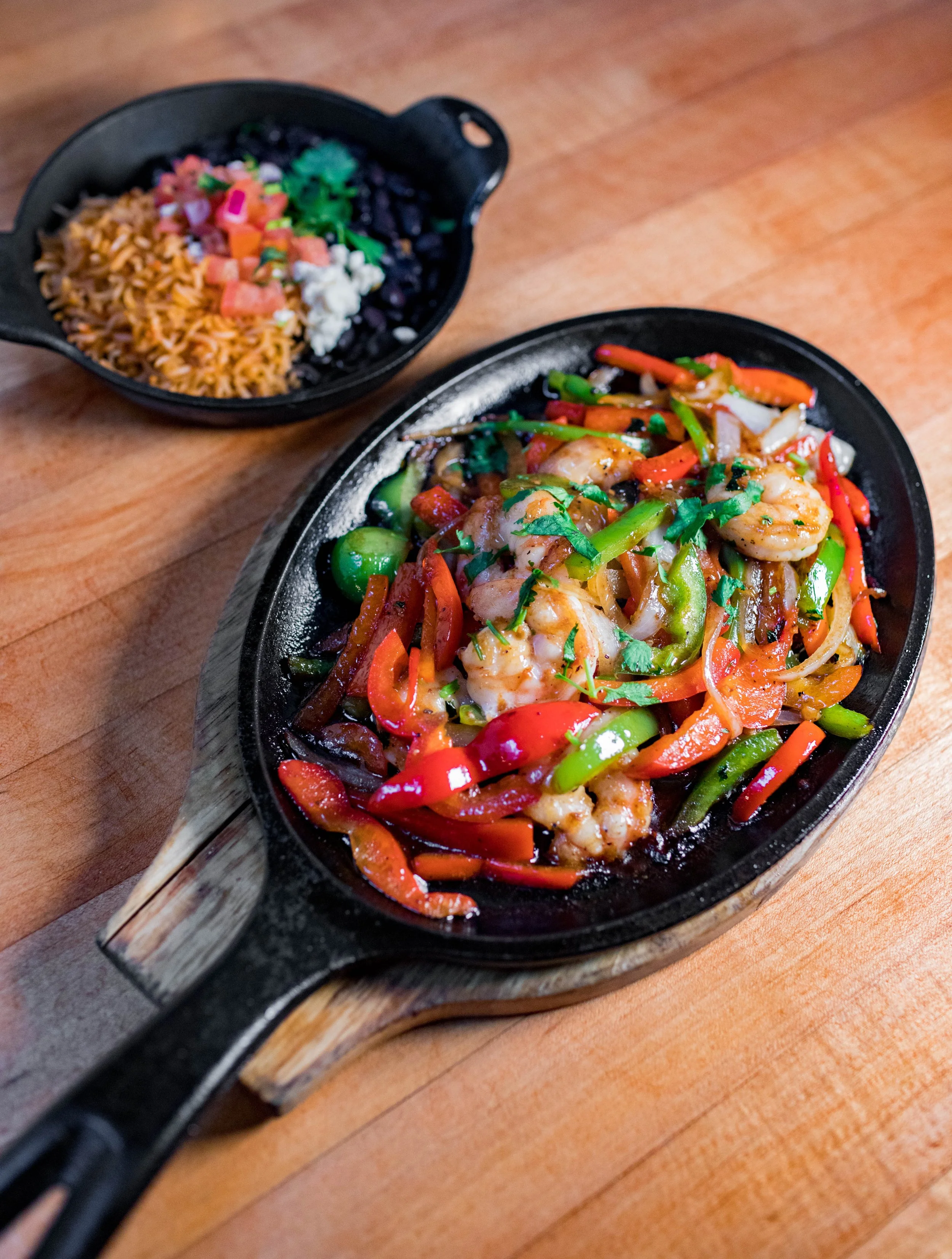 A sizzling plate of shrimp stir-fry with red and green bell peppers, onions, and cilantro, served in a cast iron skillet with a wooden handle, placed on a wooden table.