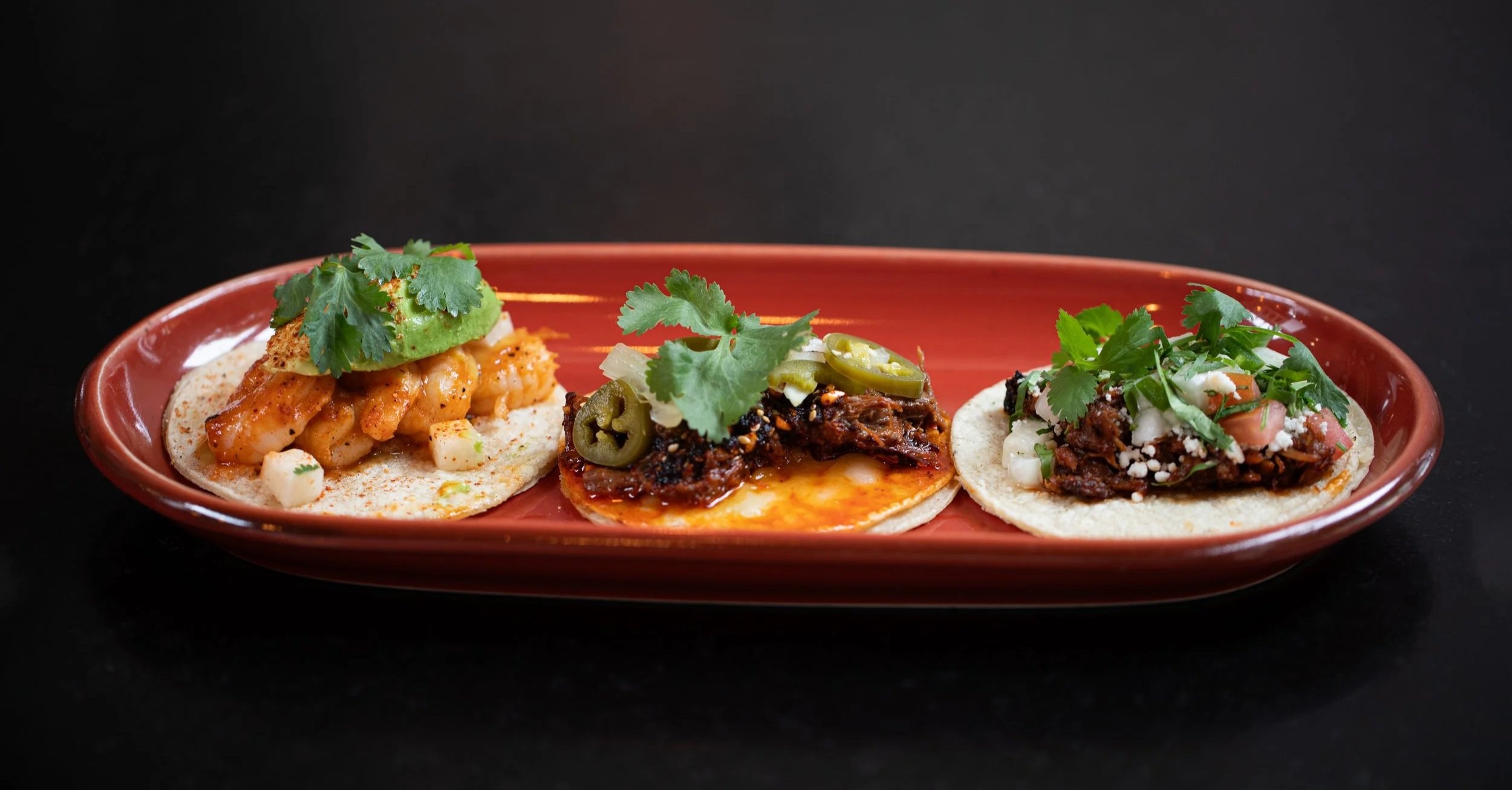 Three tacos on a red plate with cilantro garnish, each filled with different ingredients including shrimp, shredded beef with jalapenos, and shredded beef with cheese, onions, and cilantro.