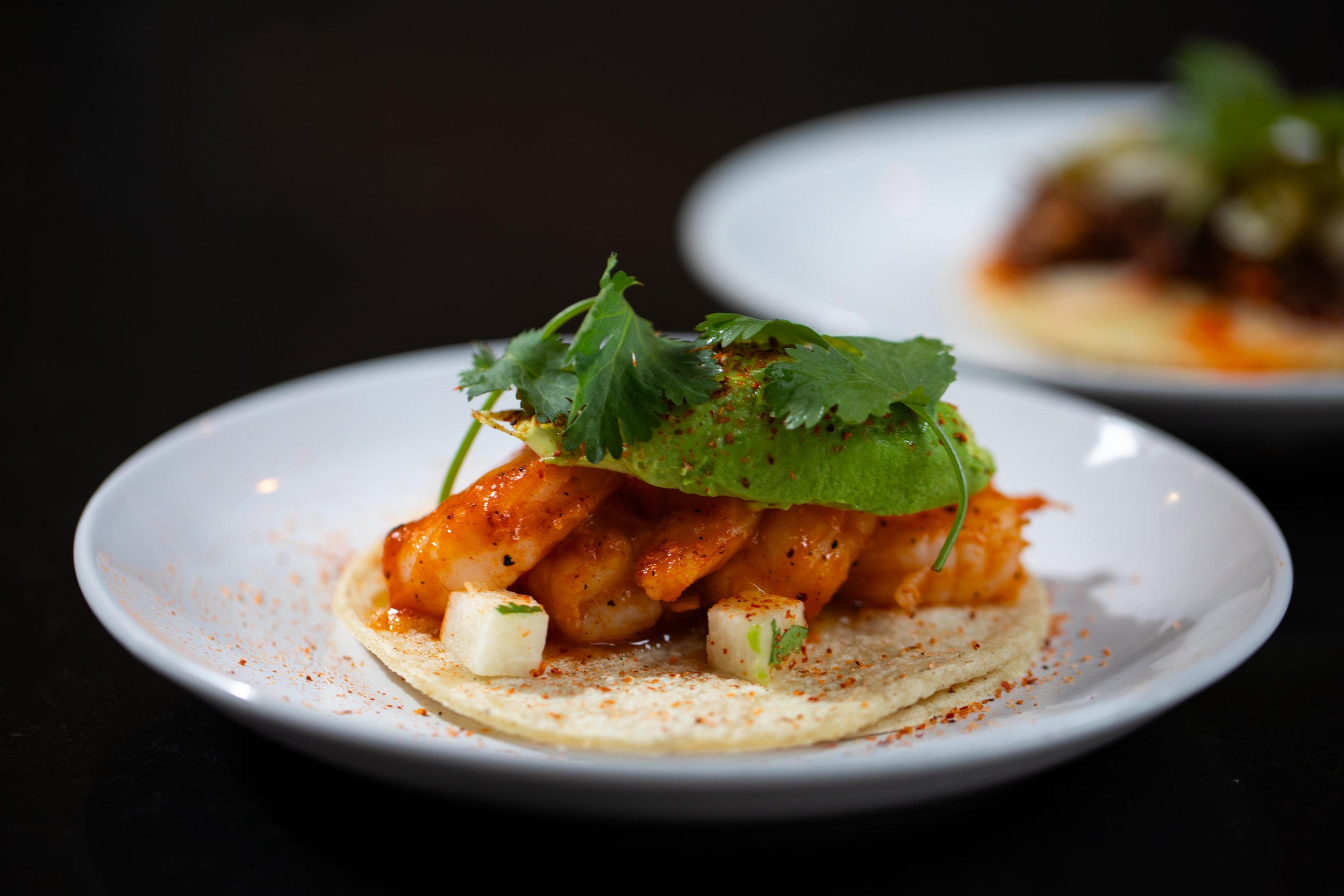 A close-up of an open-faced taco topped with cilantro, avocado, shrimp, served on a white plate against a black background.