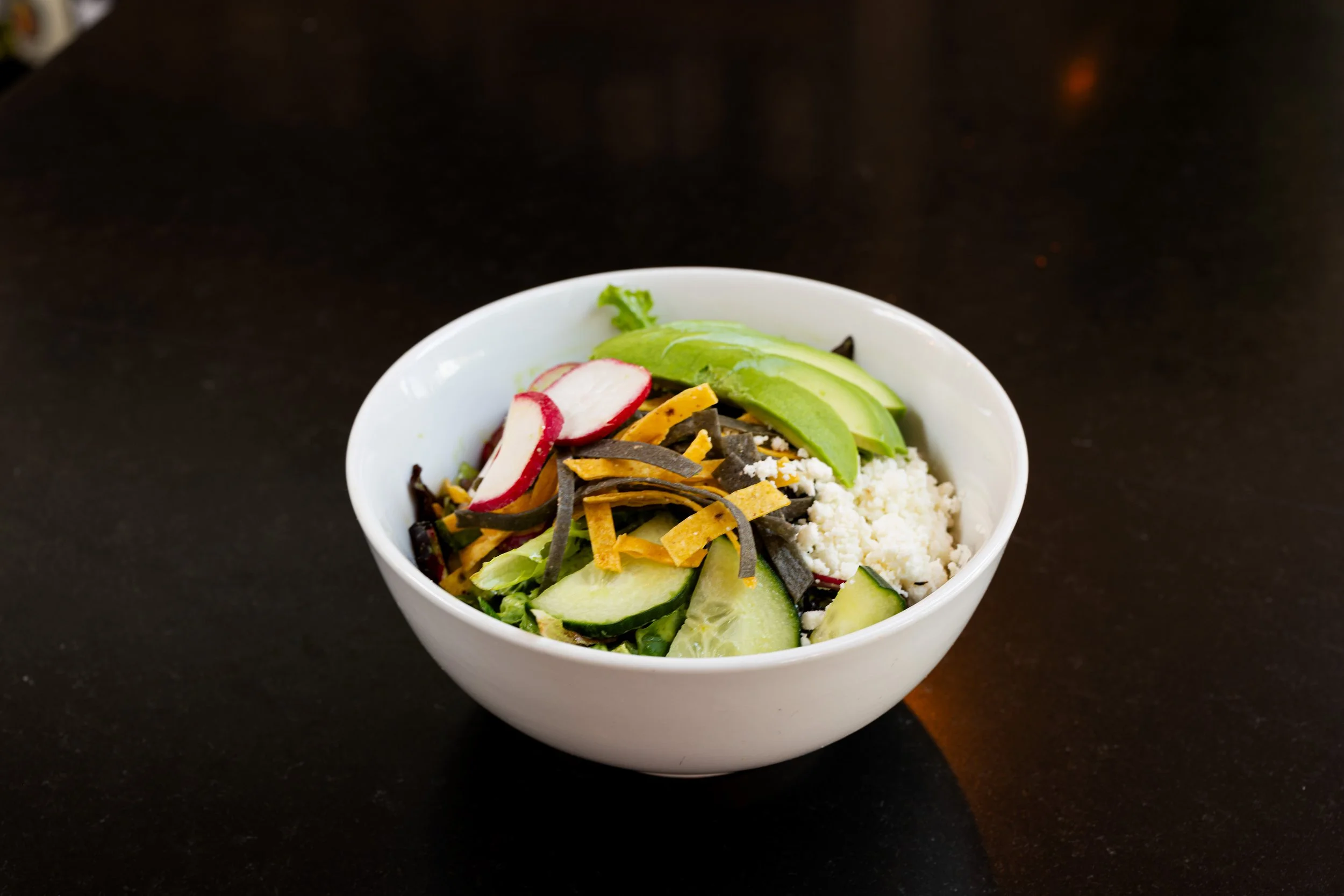 A white bowl filled with Papito Hayes' salad containing sliced radishes, avocado, cucumber, shredded cheese, black shredded seaweed, and queso, on a black bar counter