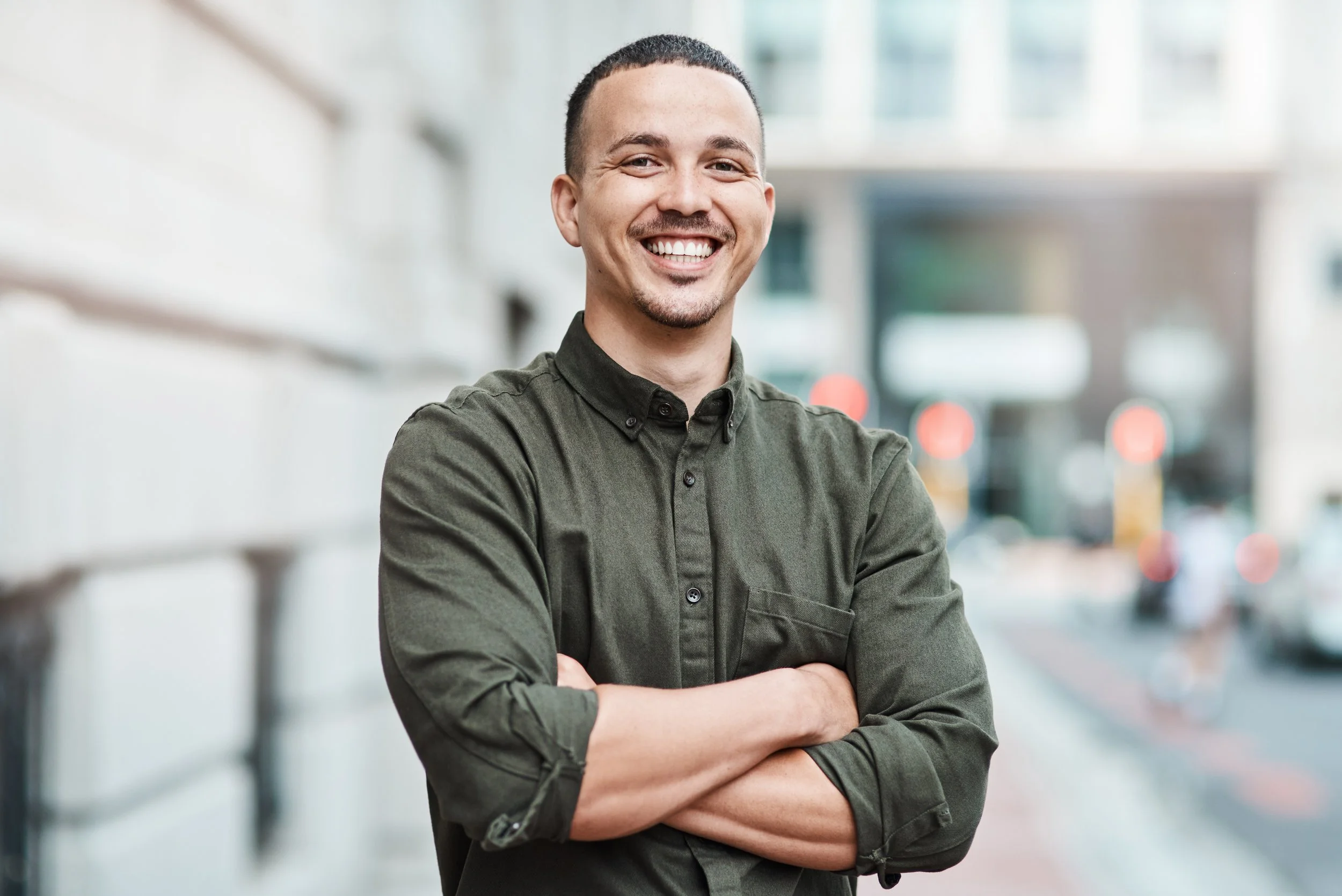 Young man with dark hair and beard smiling confidently outdoors, arms crossed, in an urban setting with blurred city background.