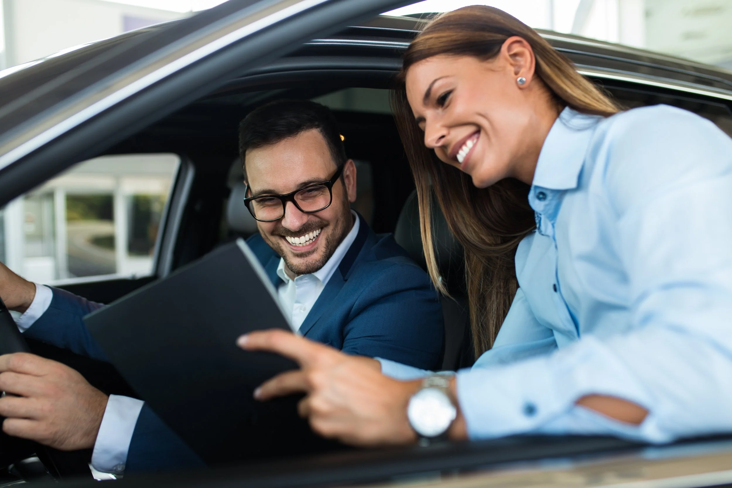 A man in a suit and glasses and a woman in a light blouse sitting inside a car, smiling and looking at a tablet together.