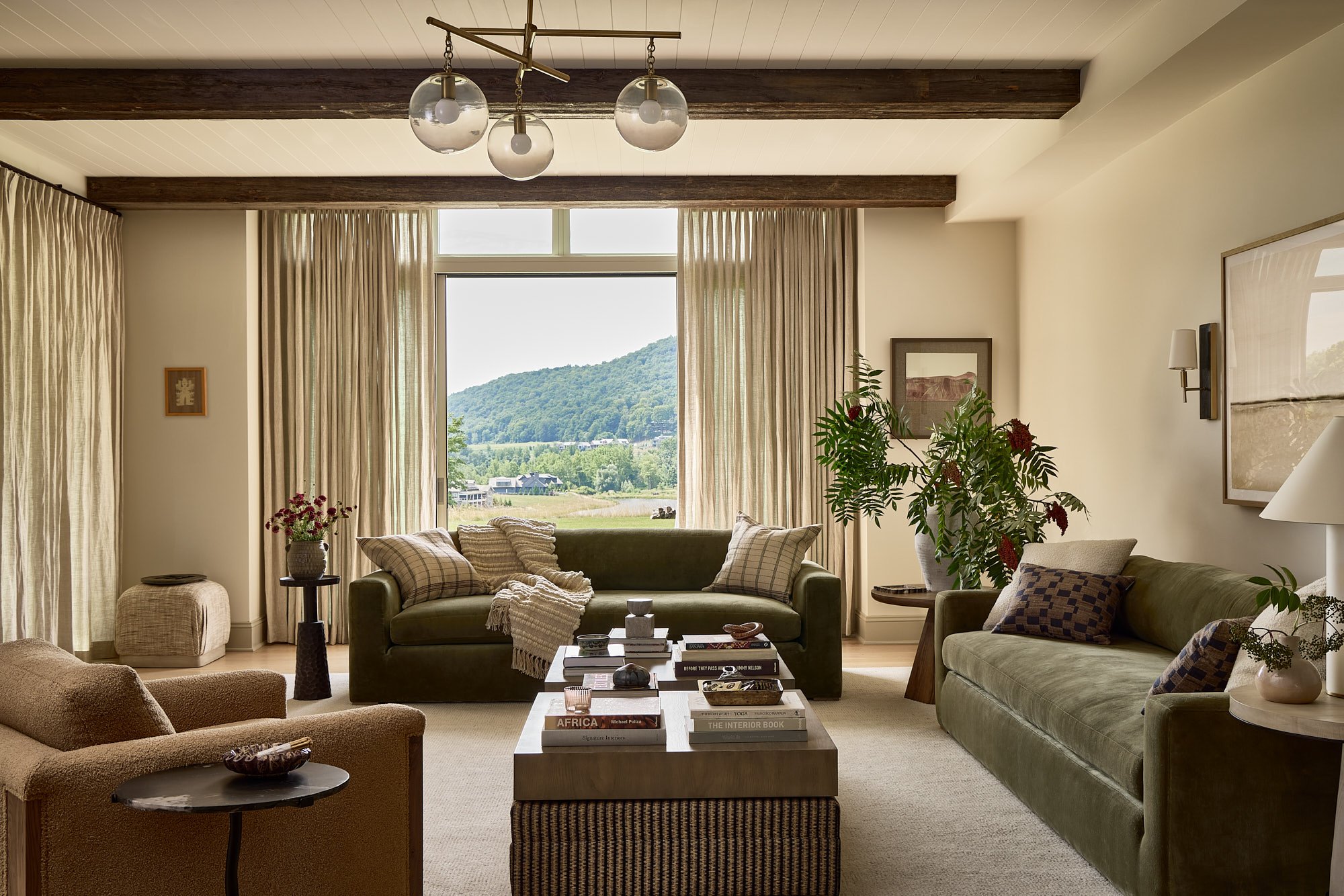 Living room with large window showing a green hill landscape, decorated with two green sofas, a beige armchair, a wooden coffee table with books, and various plants and artwork.