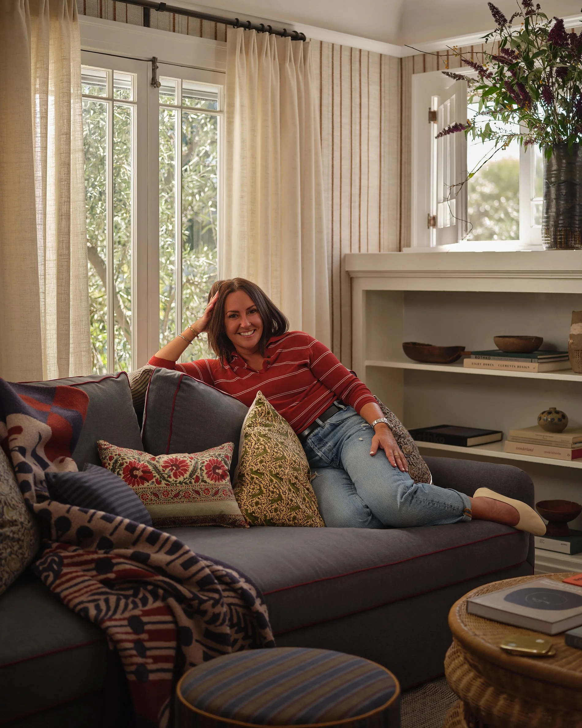 A woman relaxing on a gray sofa with colorful pillows in a cozy living room with large windows, cream curtains, and a white built-in shelf with decorative items and books.