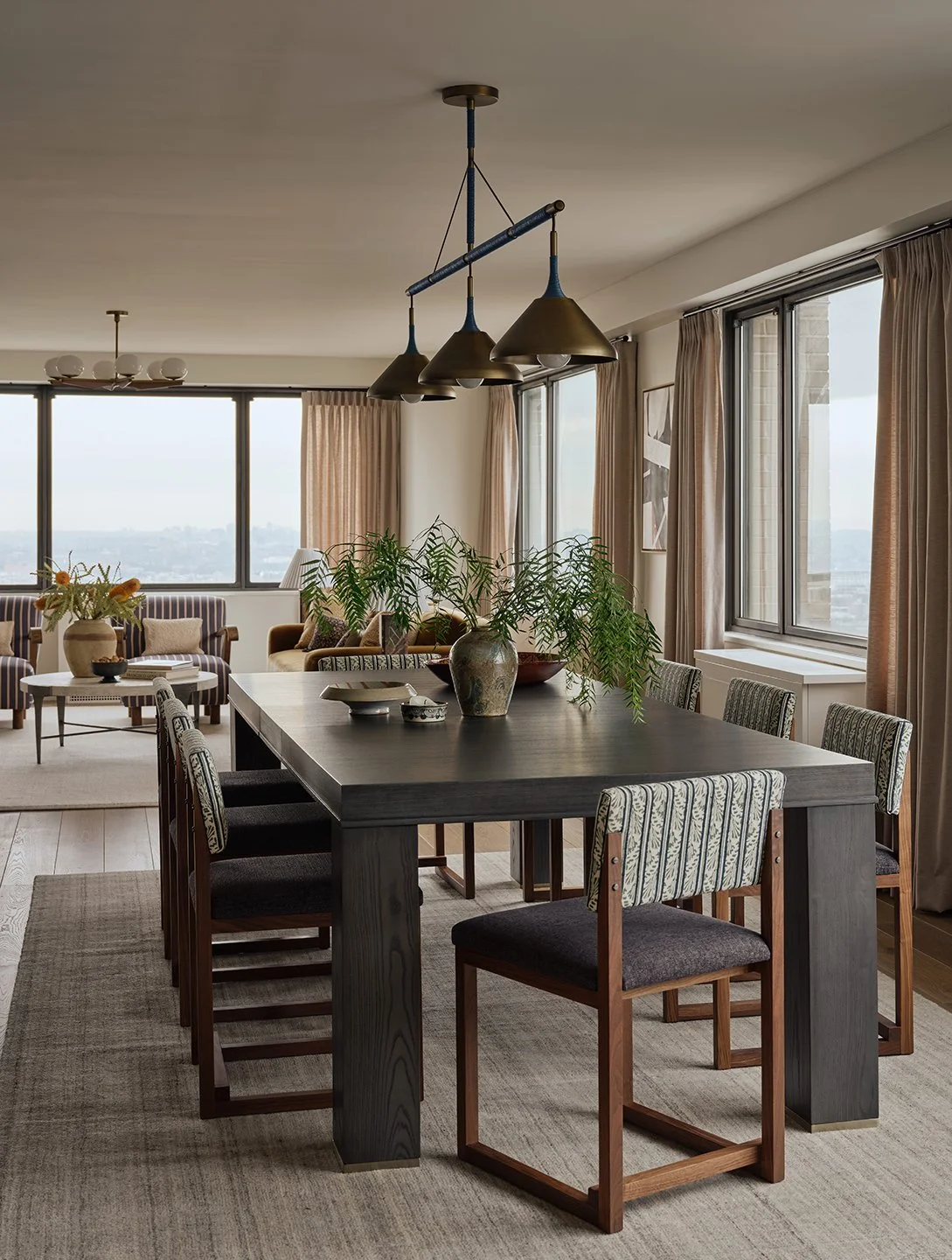 A spacious dining area with a large dark wooden table, surrounded by eight chairs with patterned upholstery, and decorated with potted plants and ceramic bowls. The room has large windows with beige curtains, light-colored hardwood flooring, and modern lighting fixtures hanging from the ceiling.