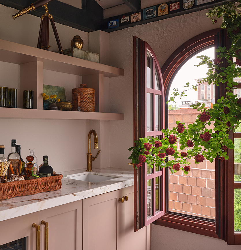 A cozy kitchen corner with a window open to outside, decorated with a flowering plant in a window box, a marble countertop with bottles and a basket, and a white cabinet with brass handles.