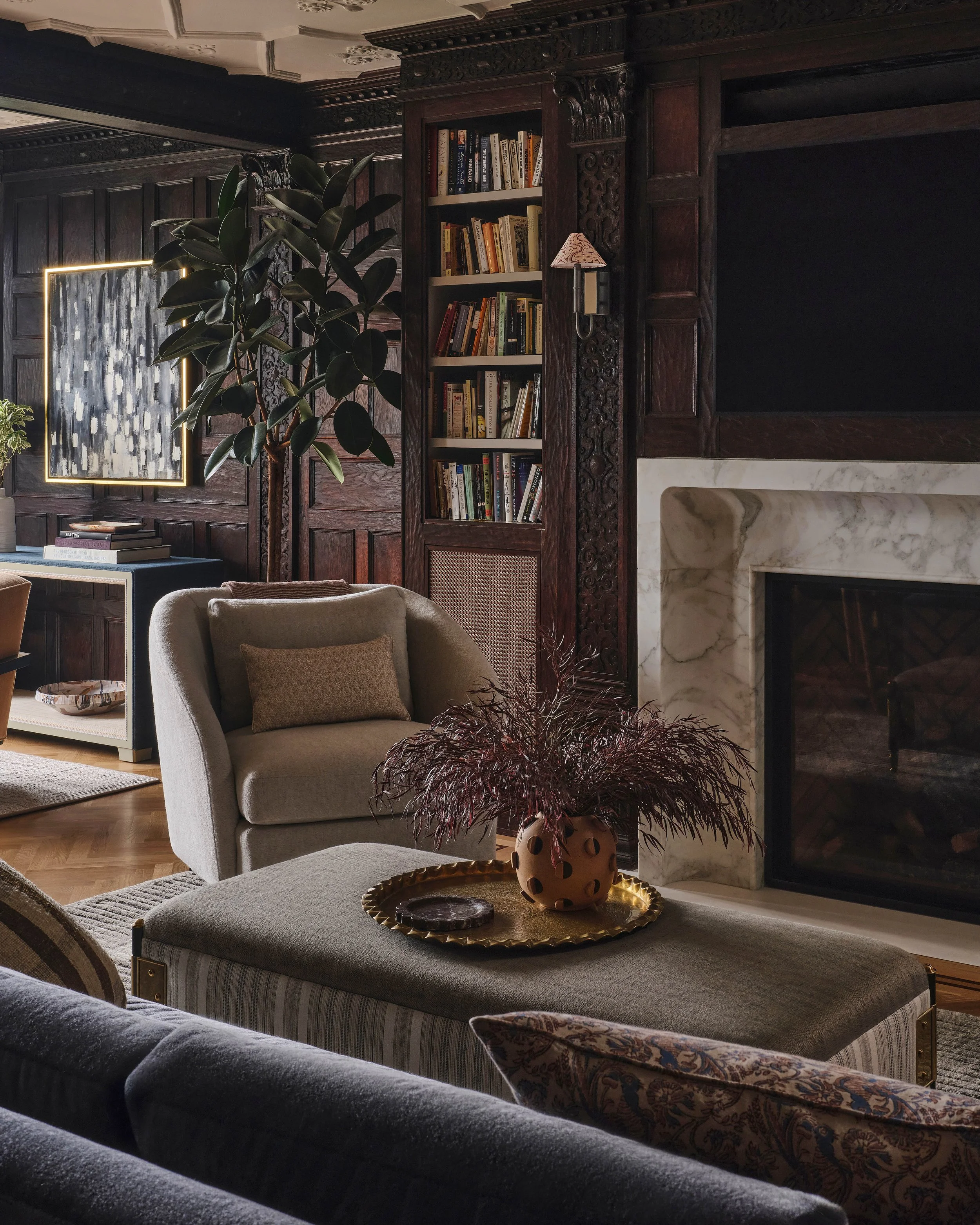 A living room featuring dark wooden paneled walls, a fireplace with a marble surround, a tall built-in bookshelf filled with books, a beige armchair with a small pillow, a potted plant, a large abstract painting, and a coffee table with a decorative vase and tray.