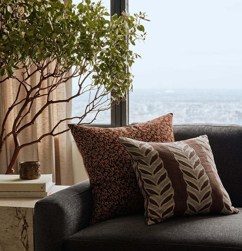 Living room with a dark gray sofa decorated with two patterned throw pillows, next to a side table with books and a decorative cup. There is a large indoor plant and a window showing a cityscape in the background.